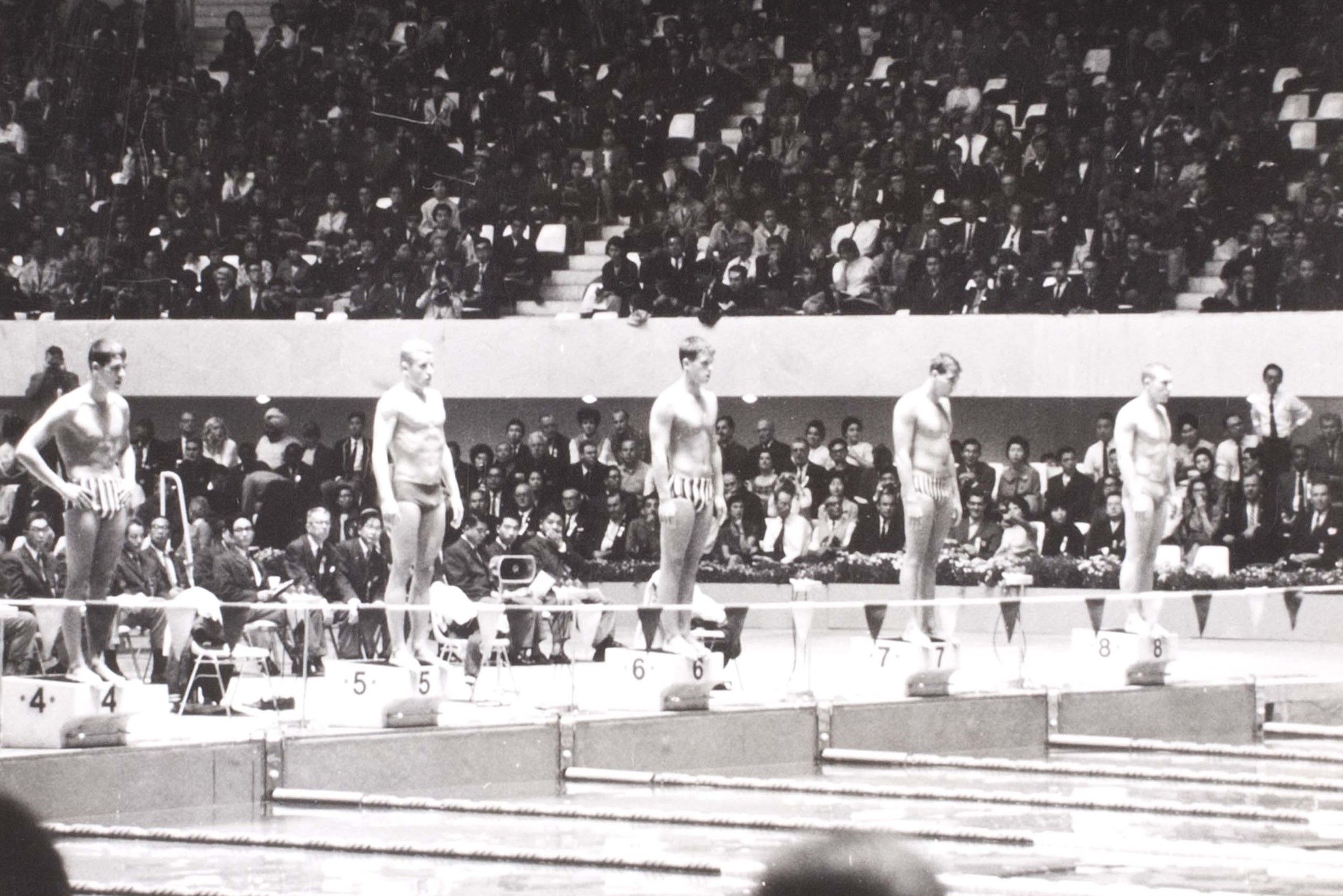 Black and White photo showing a line of 5 swimmers at one end of a pool preparing for a race with a crown behind them.