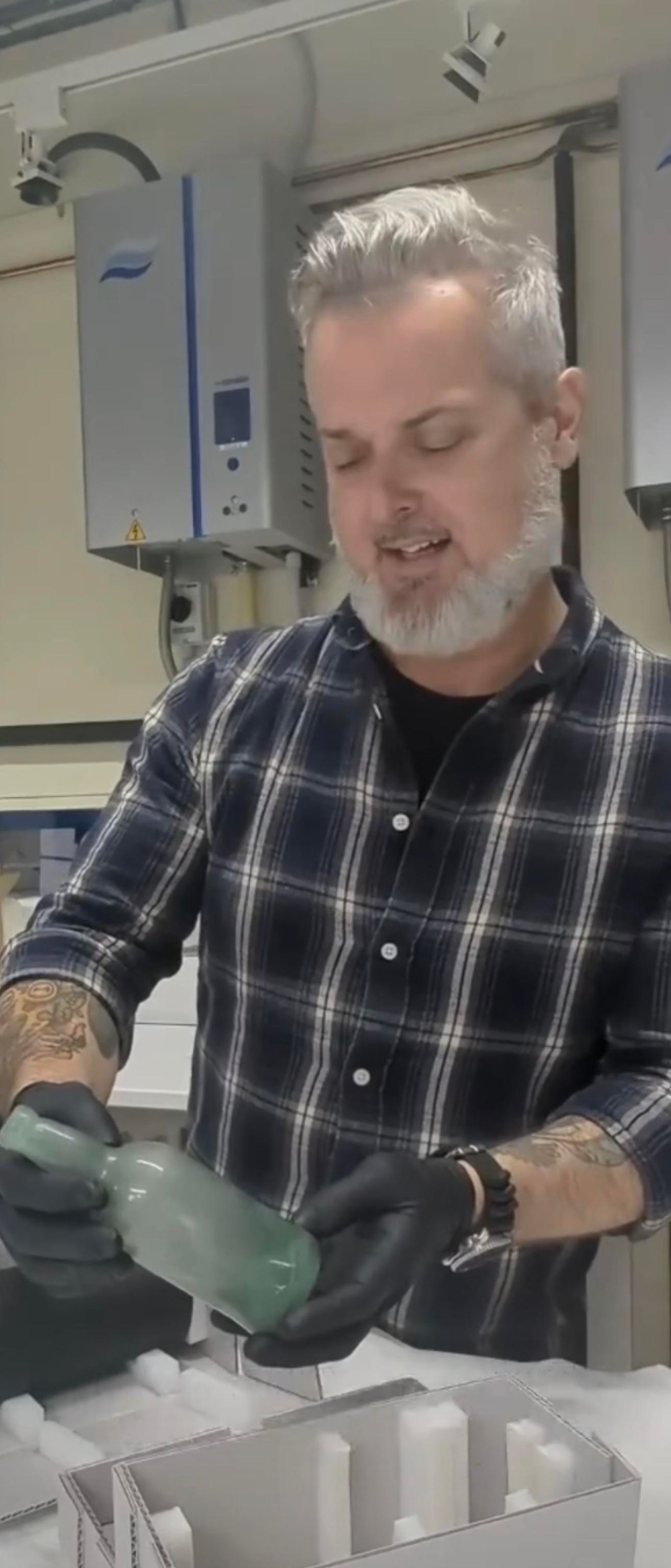 Photo of a man wearing a check shirt and gloves and holding a glass bottle.