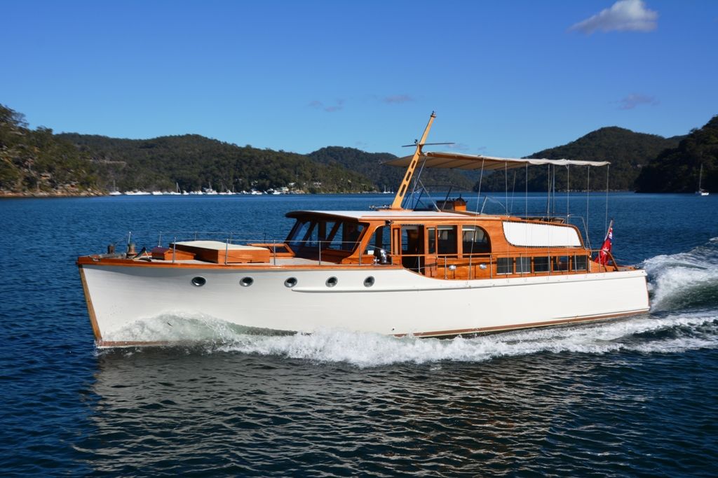 Photo of a sleek, white and timber boat on blue water, with green hills and a clear blue sky behind it.