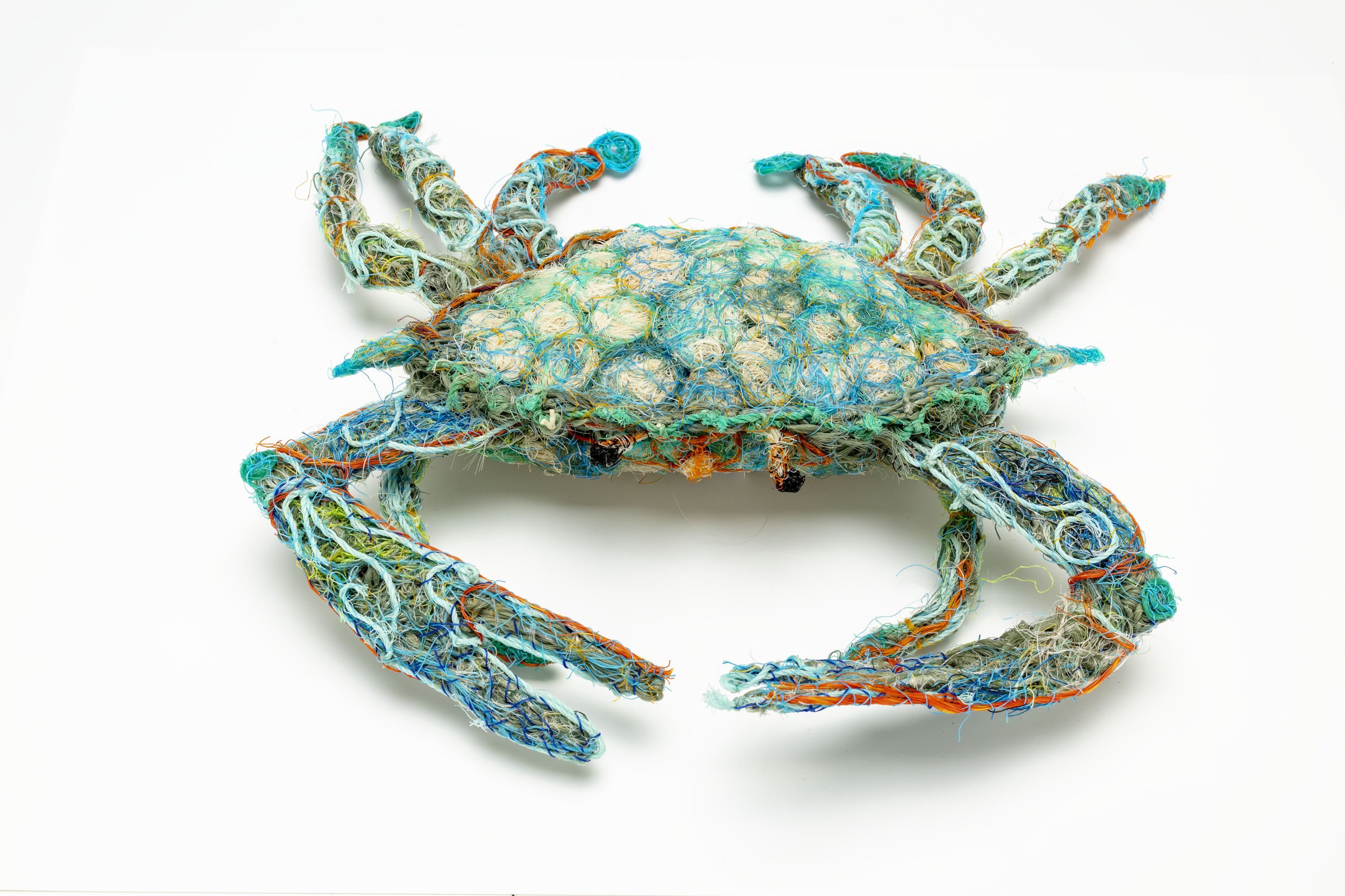 a soft sculpture depicting a crab made from discarded nets at sea from different colours, including dark and light blue, orange, green, aqua, oatmeal, clear and black. The sculpture’s surface has different patterns created by the nets which are woven tight so the filling of further nets is tightly secured.