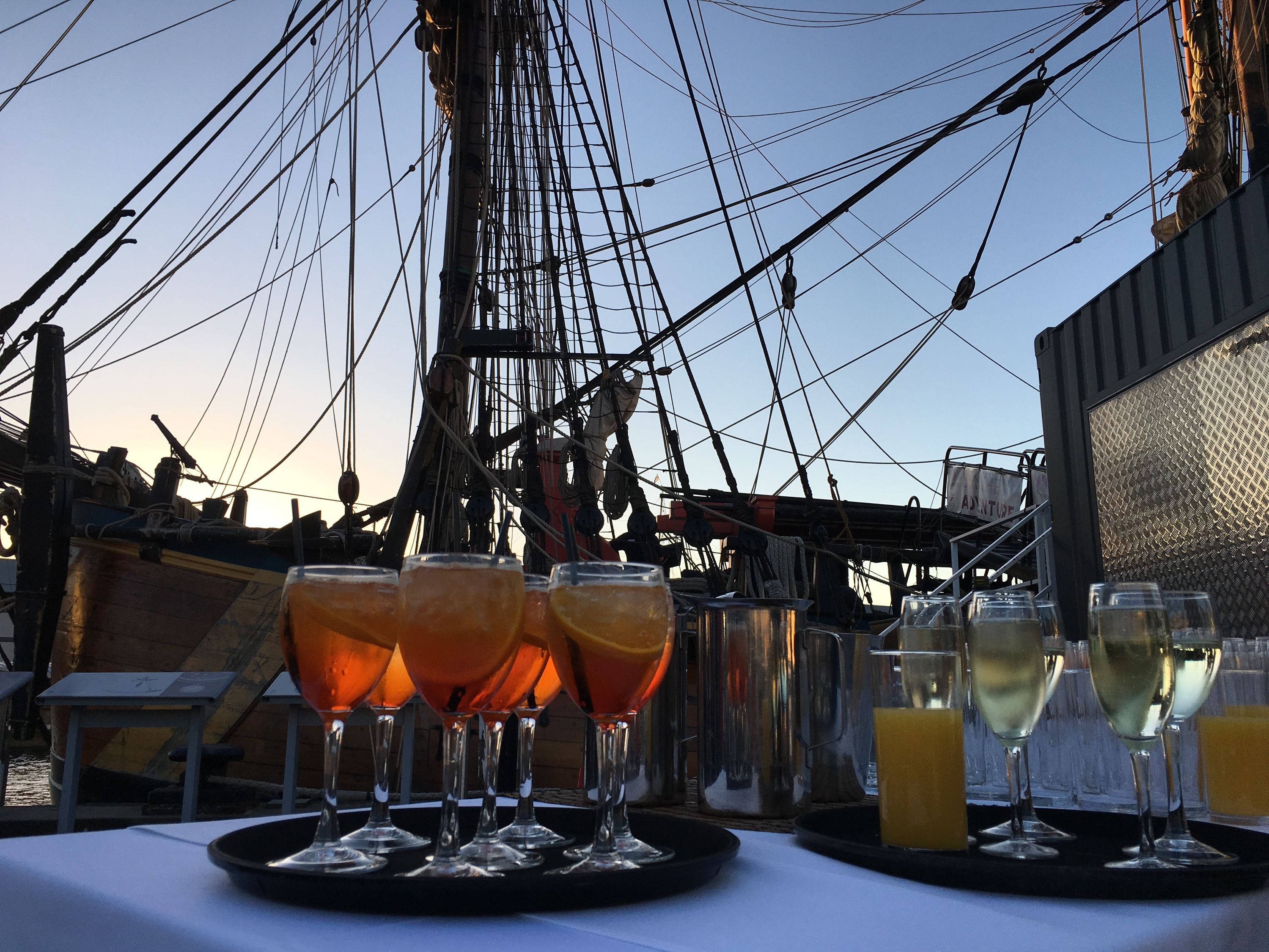 Photo of cocktails on a table in front of tall ship masts. 