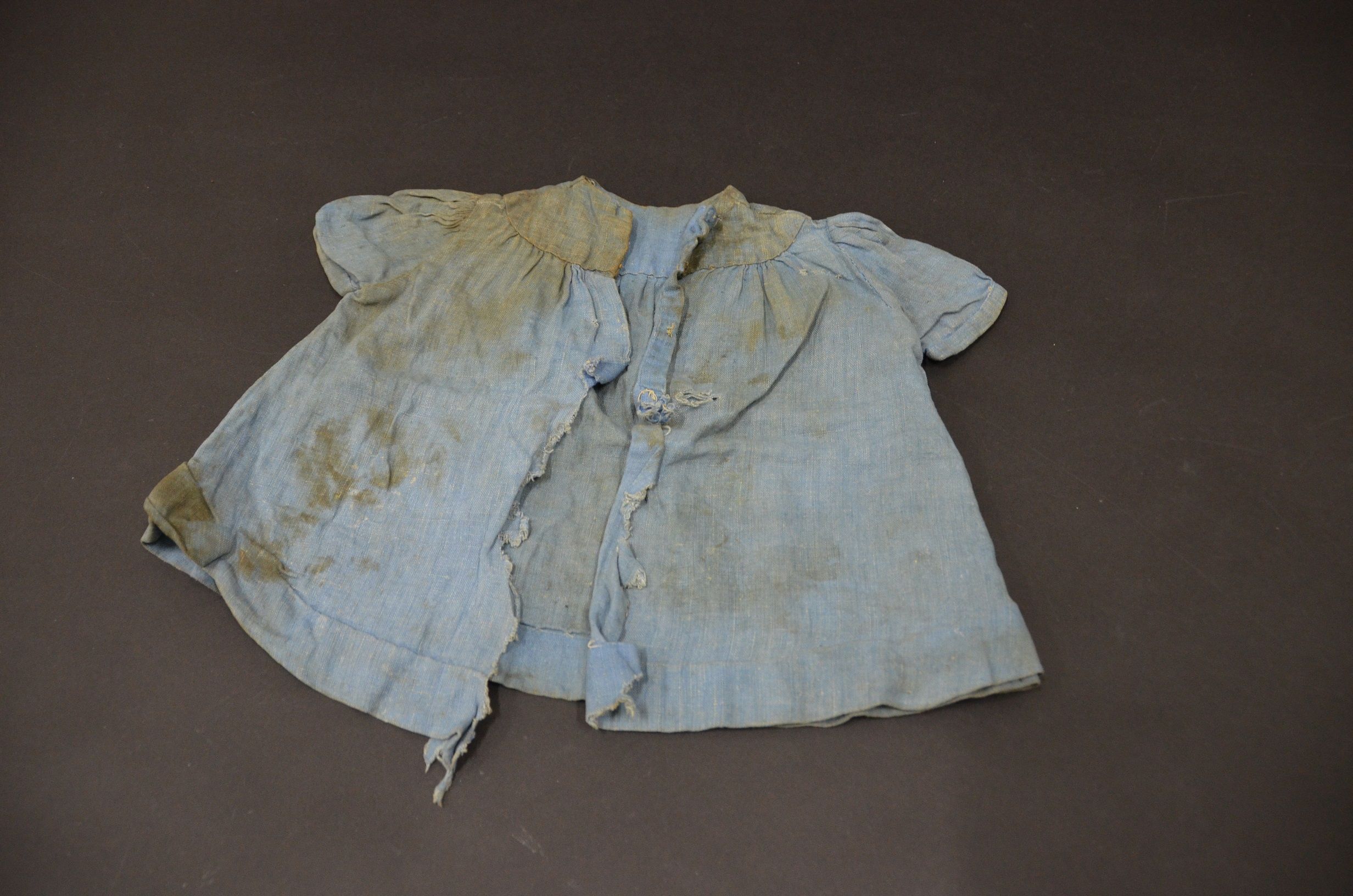 Photo of a light coloured, child's dress with yellow brown stains. The fabric is ripped around the front buttons. 