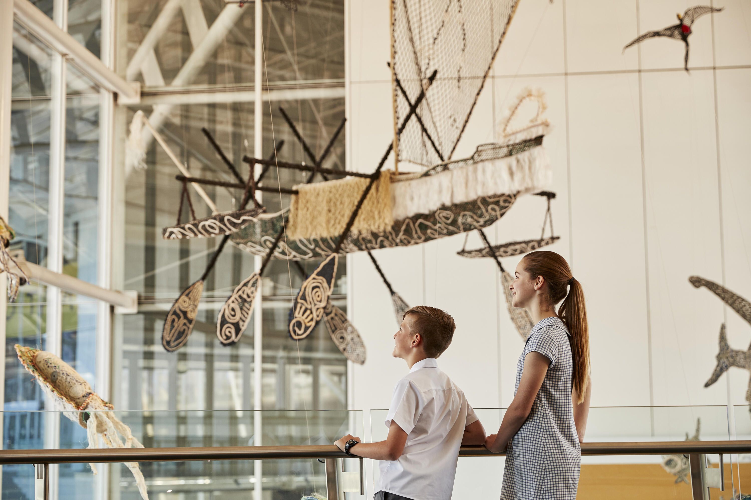 2 School kids exploring the museum, stop to look at the ghost net sculptures suspended in the foyer.