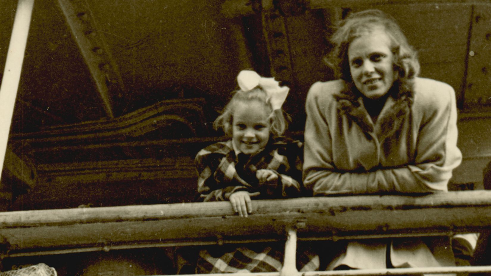 Black and white photo on yellowed paper showing A young girl and a woman are pictured standing behind the railing on the deck of a ship. The girl wears a large bow in her hair.