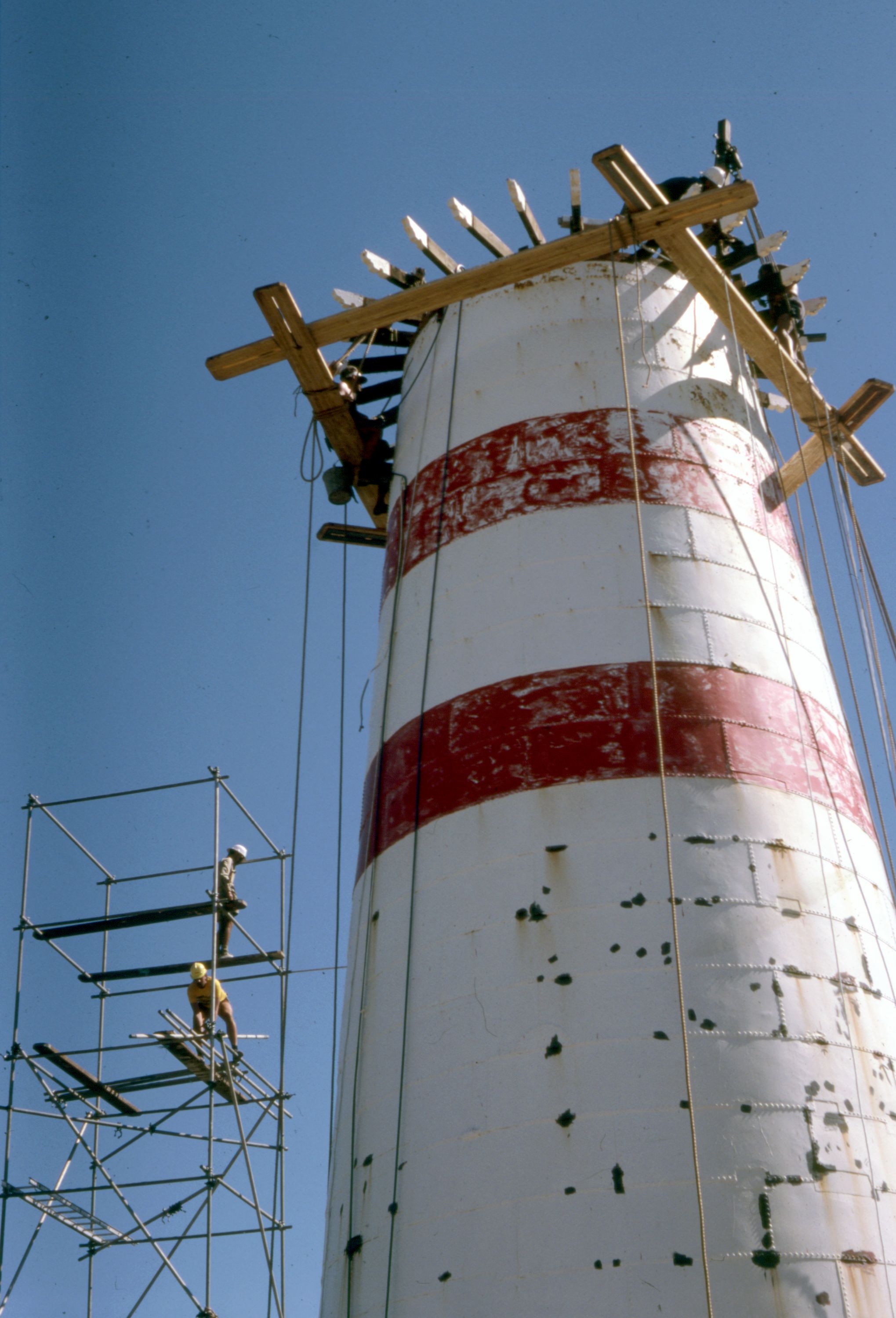 Photograph looking up at a partially dismantled lighthouse which is white, with red stripes, with a blue sky behind.  