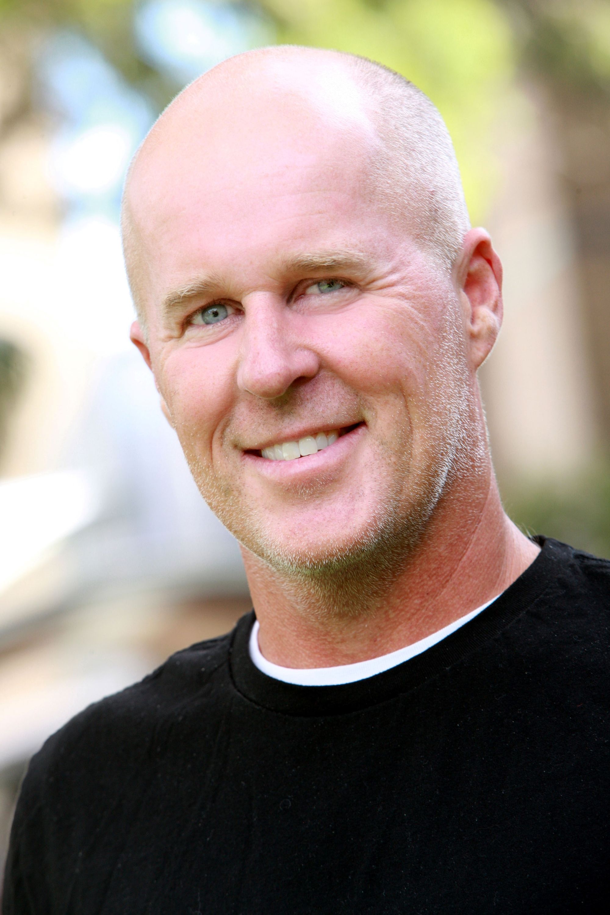 Photo of a man, smiling with light skin, a bald head and black top.