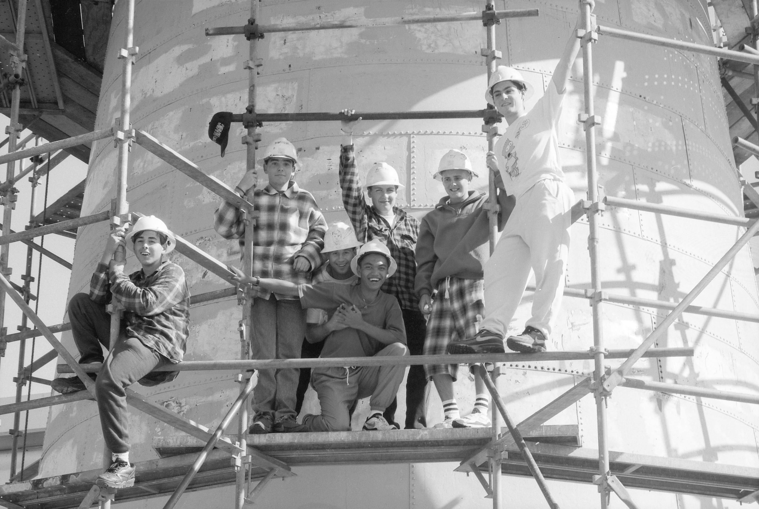 Black and white photo of a group of young men together on scaffolding around a lighthouse. 