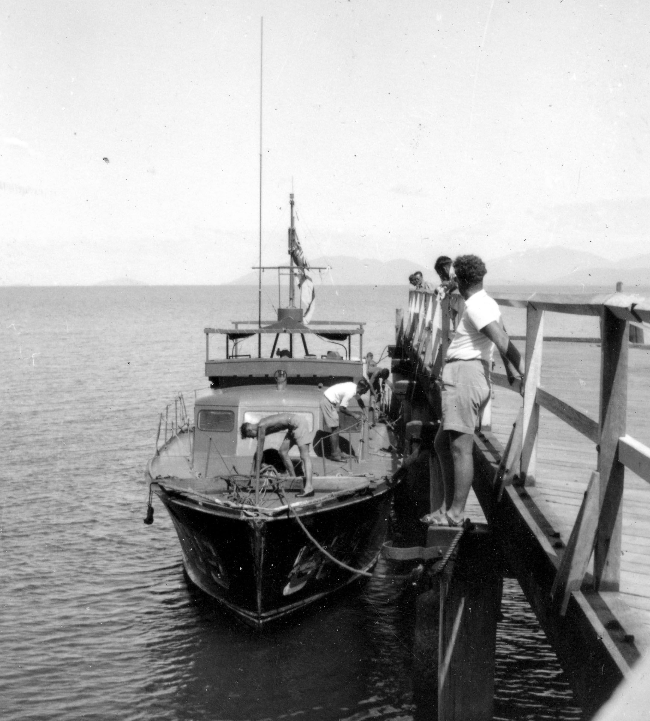 Black and white photo of a boat tied up to a wharf, with people standing on the wharf.