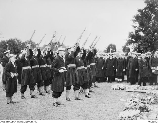 Black and white photo showing a group of men, standing in rows at a burial. They are all in dark coats, and 2 rows have rifles pointed to the sky in a salute.