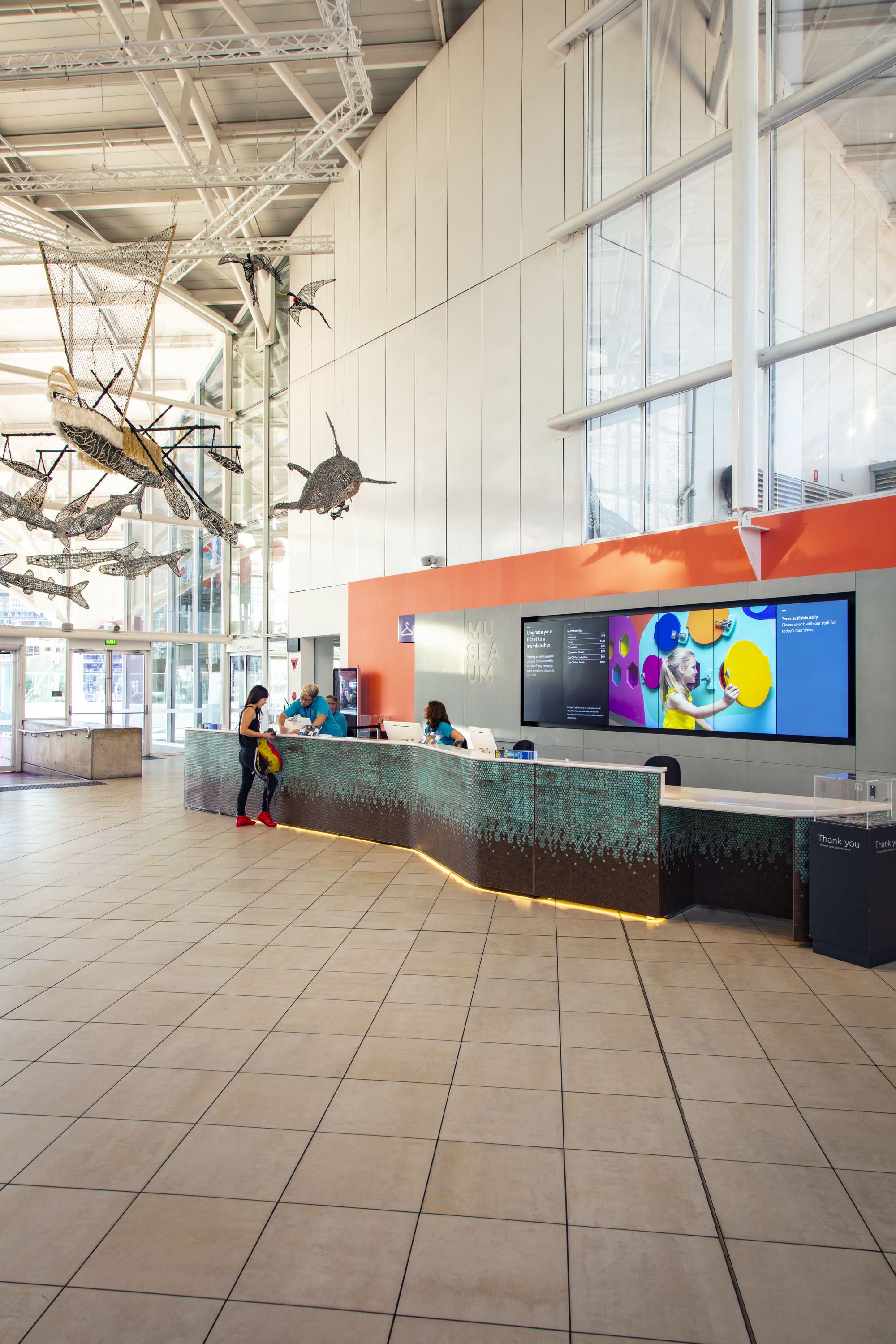 Photo showing the museum foyer near the front desk, with a visitor and museum staff. The Ghost Net sculptures appear overhead.