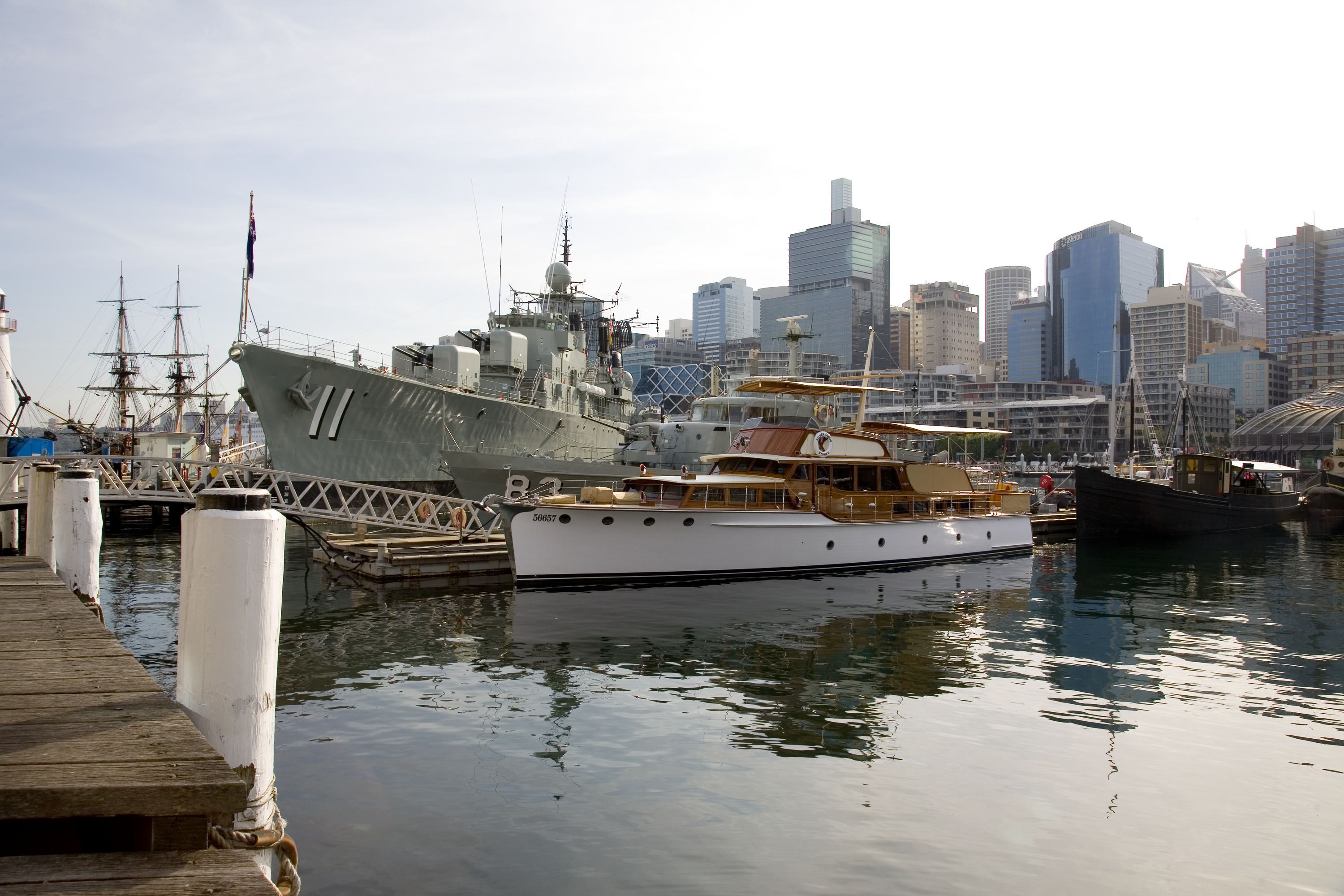 Photograph showing a wooden boat with white hull at the museum wharf, with a navy ship behind it. 