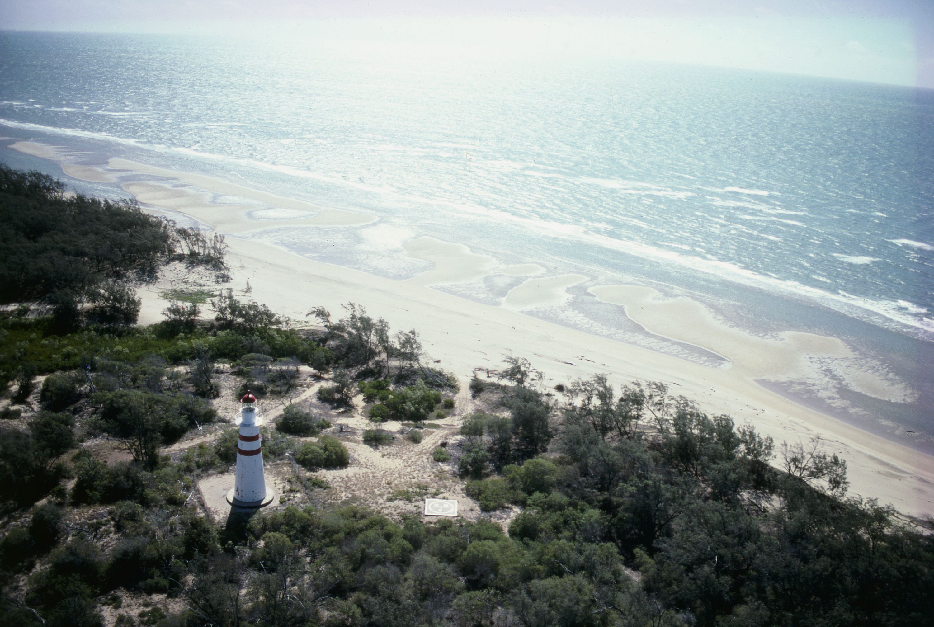 Aerial photograph of a beach and scrub, with a white lighthouse in the bottom left corner.