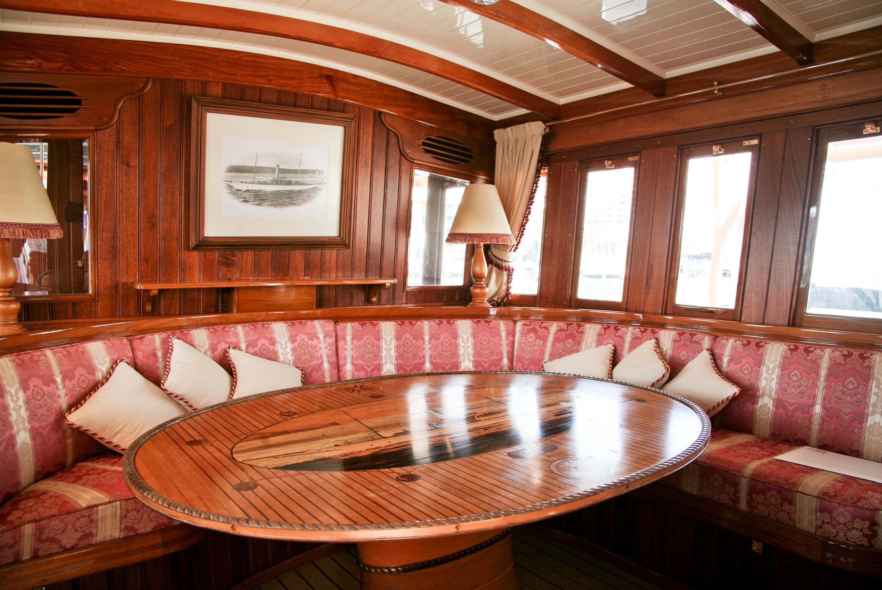 Photo of a wooden panelled cabin on a edwardian period steam yacht. There is a wooden table and bench seating with red and cream upholstery, and a black and white photo handing on the wall. 