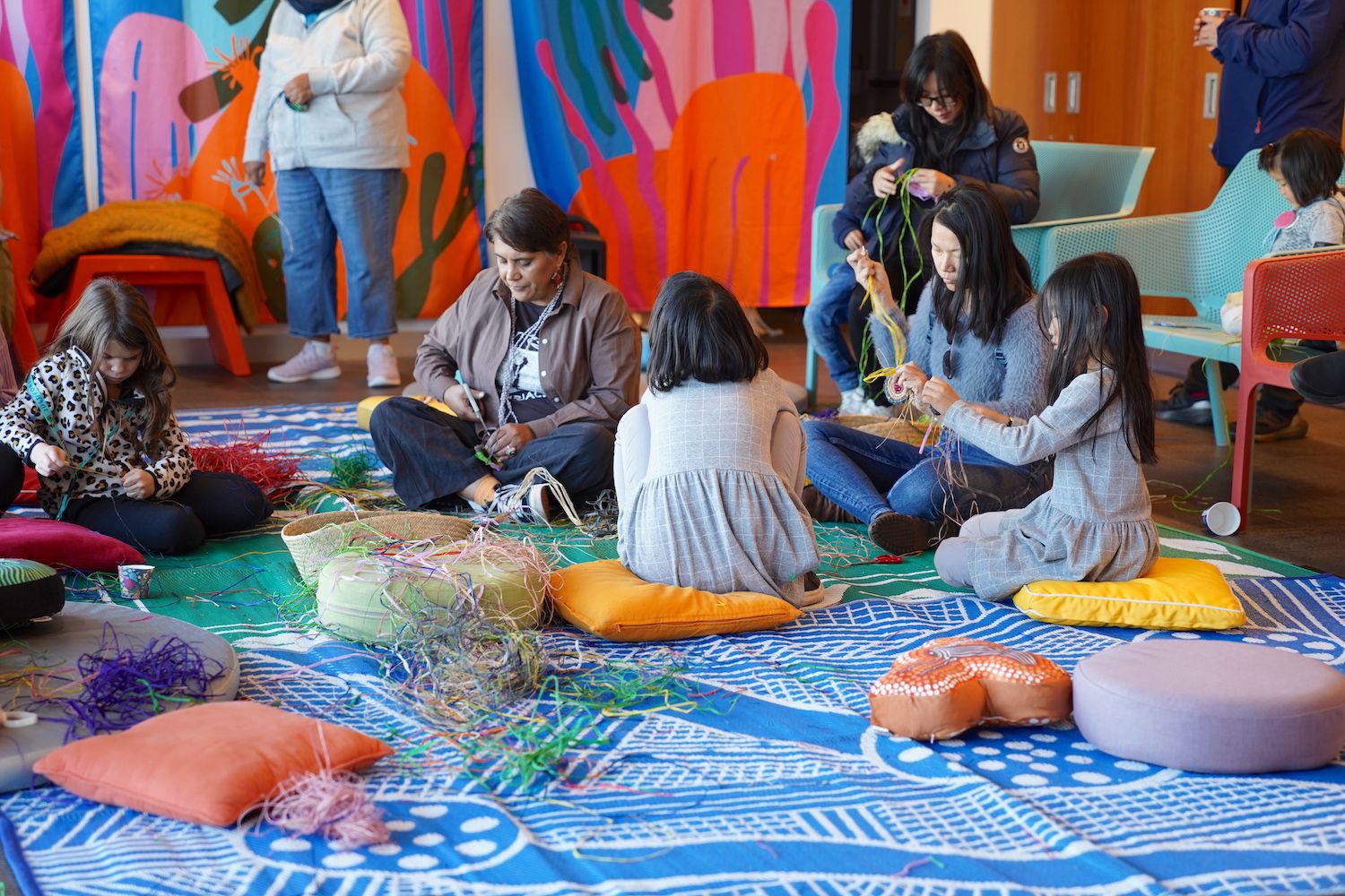 Photo of a group of children and adults sitting on cushions and weaving with twine.