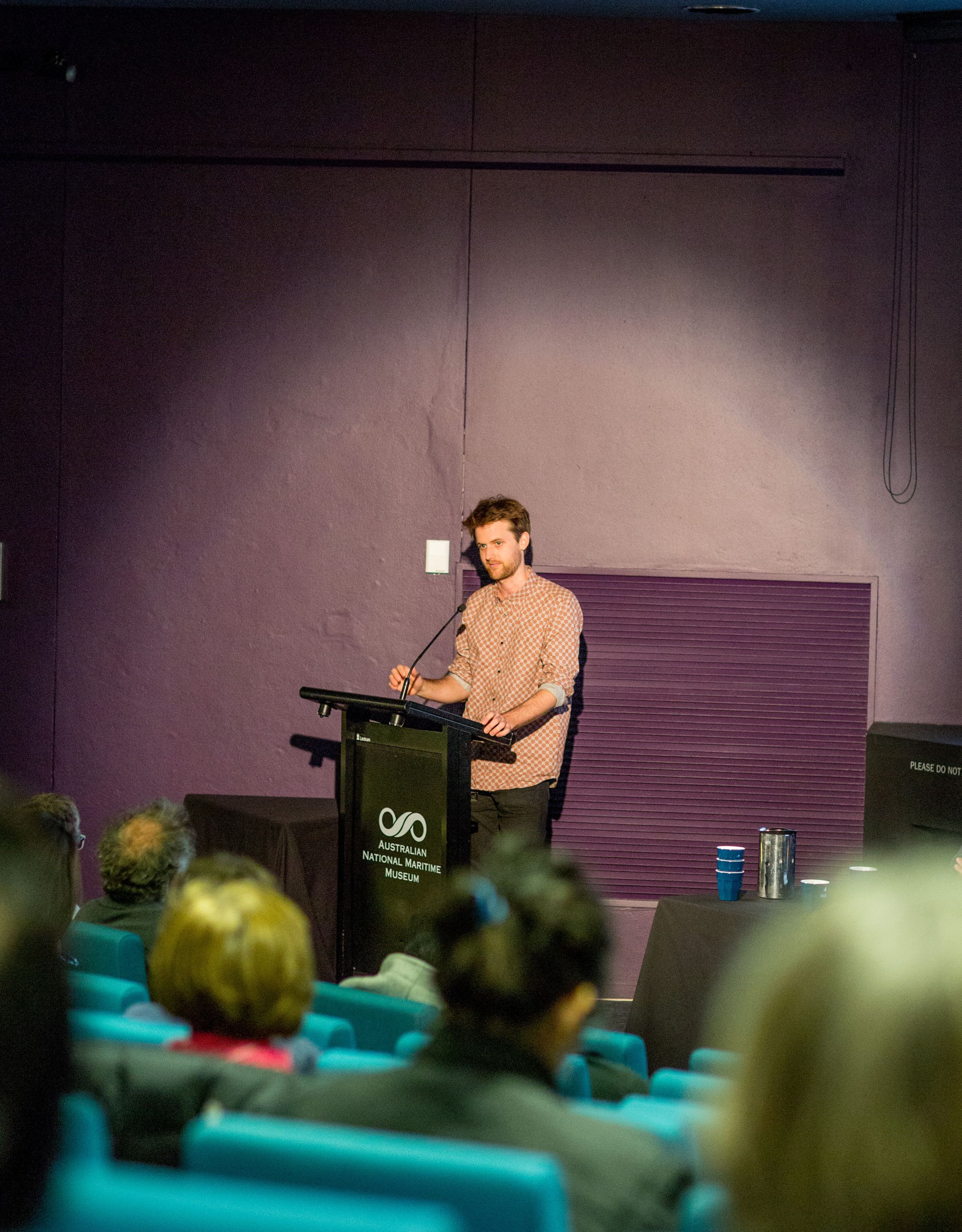 Male presenter in a spotlight, standing at a podium in front of a purple wall. 