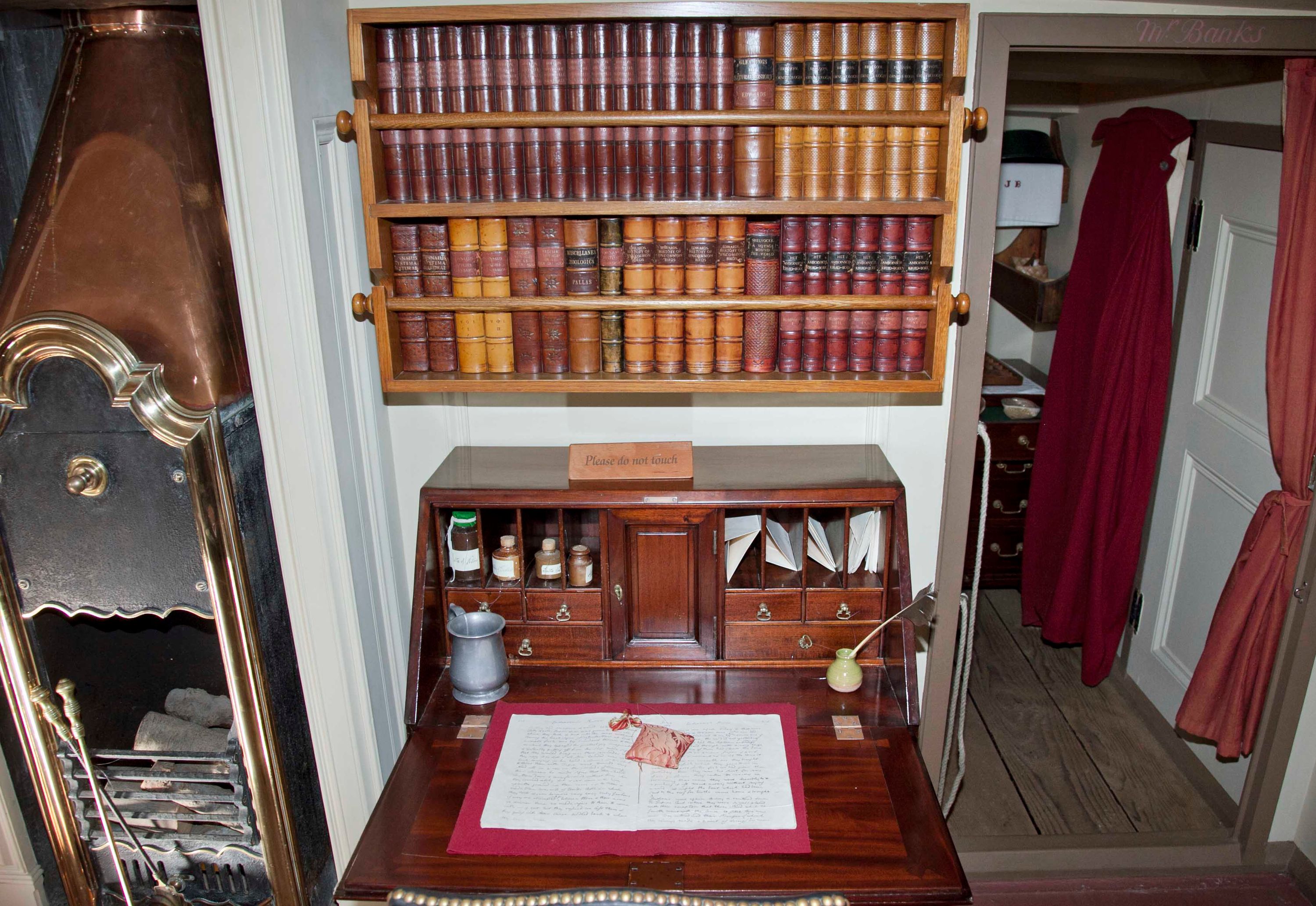 Photo of a small, wooden folding desk and bookshelf in a cabin on a wooden tall ship.
