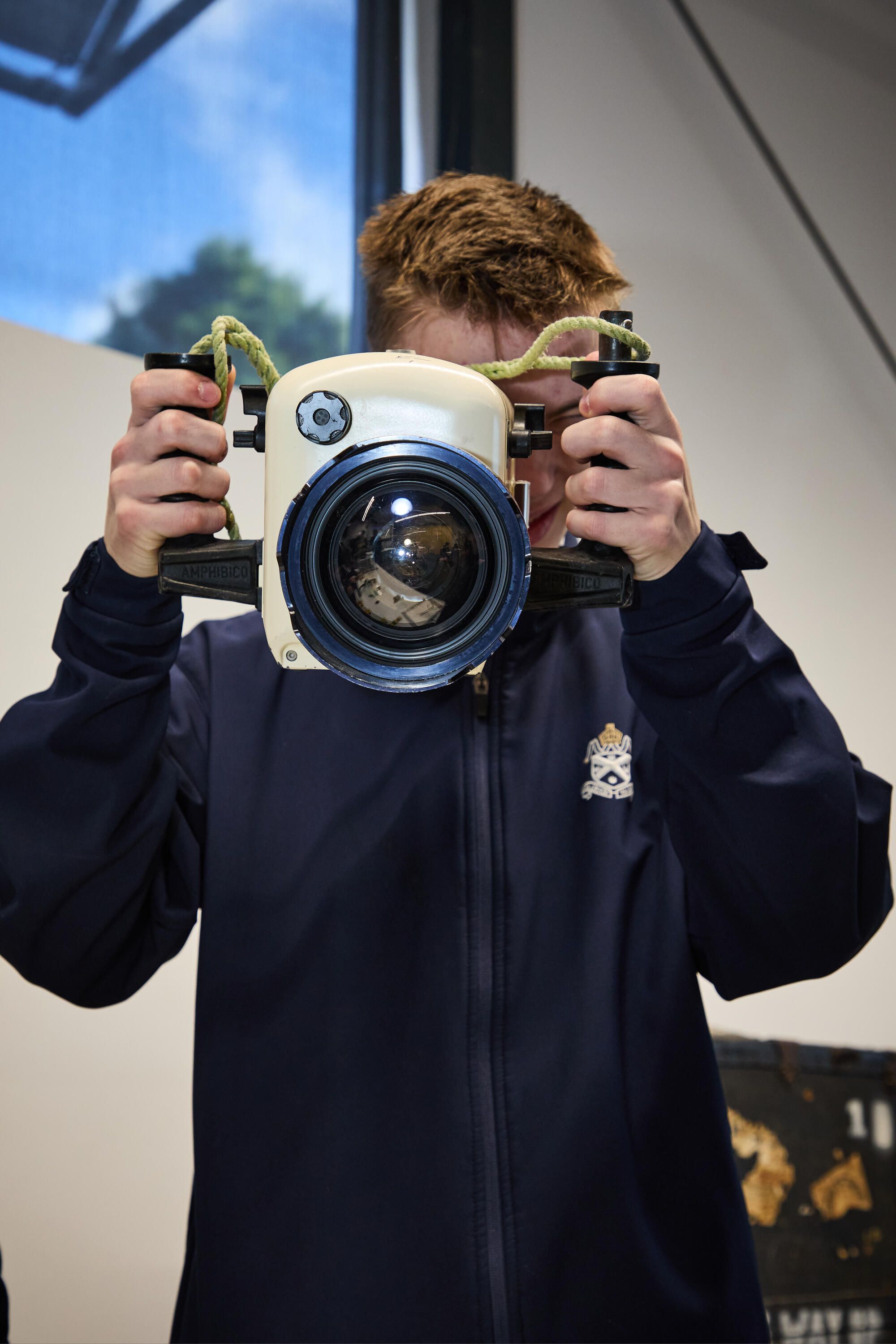 Photo showing a teen boy in a navy jacket holding a large underwater camera.