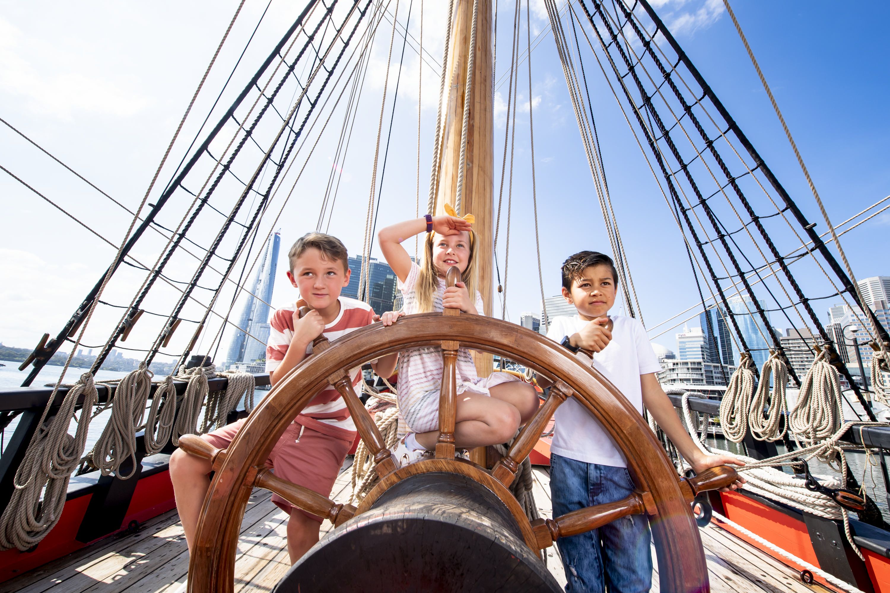 Photo showing two boys and a girl explore on board a tall ship and pose behind the ship's wheel.