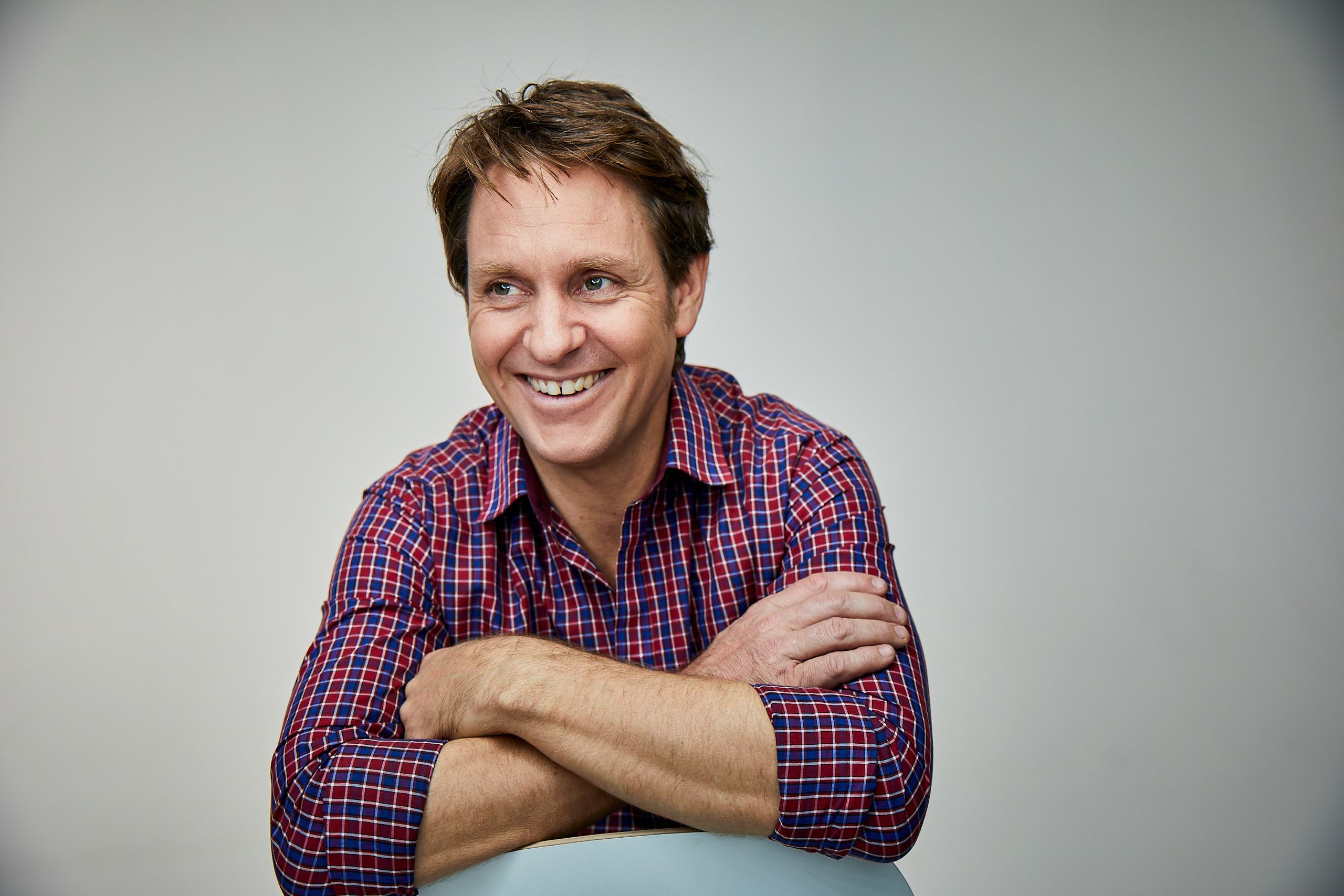 Photo of a man smiling, with short brown hair wearing a red and blue checked shirt.