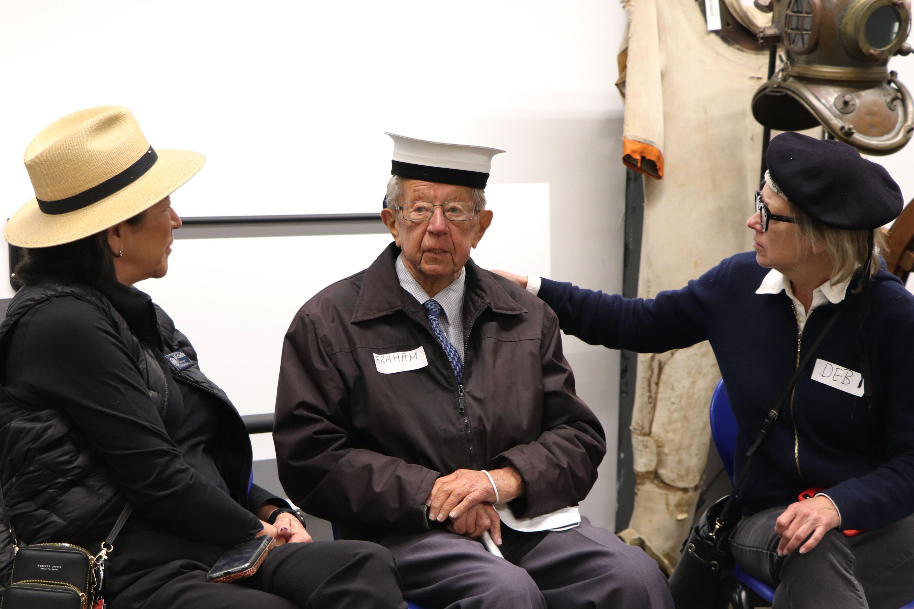 Two ladies sitting either side of an older gentleman wearing a sailors hat.