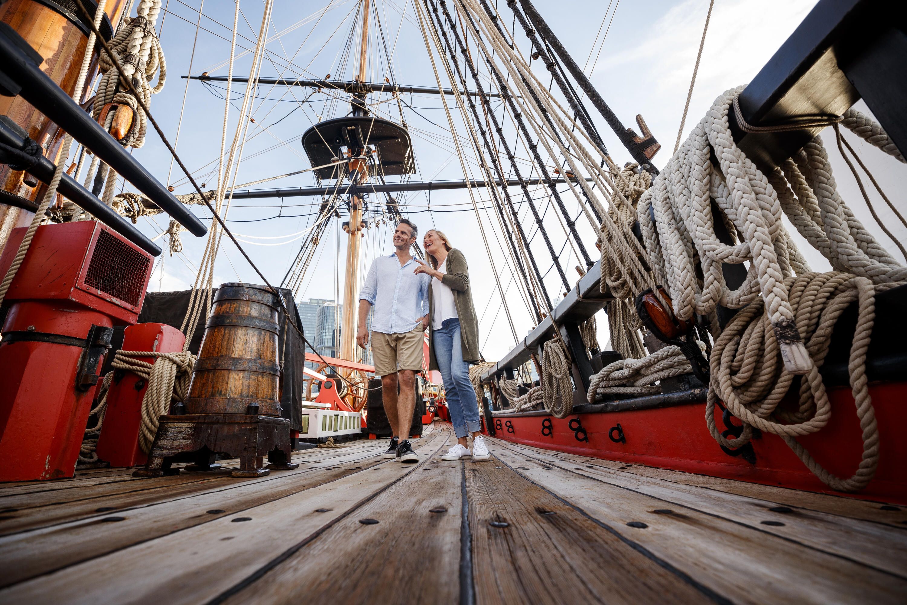 Photo taken from a low angle depicting a couple strolling along the upper deck of a wooden tall ship