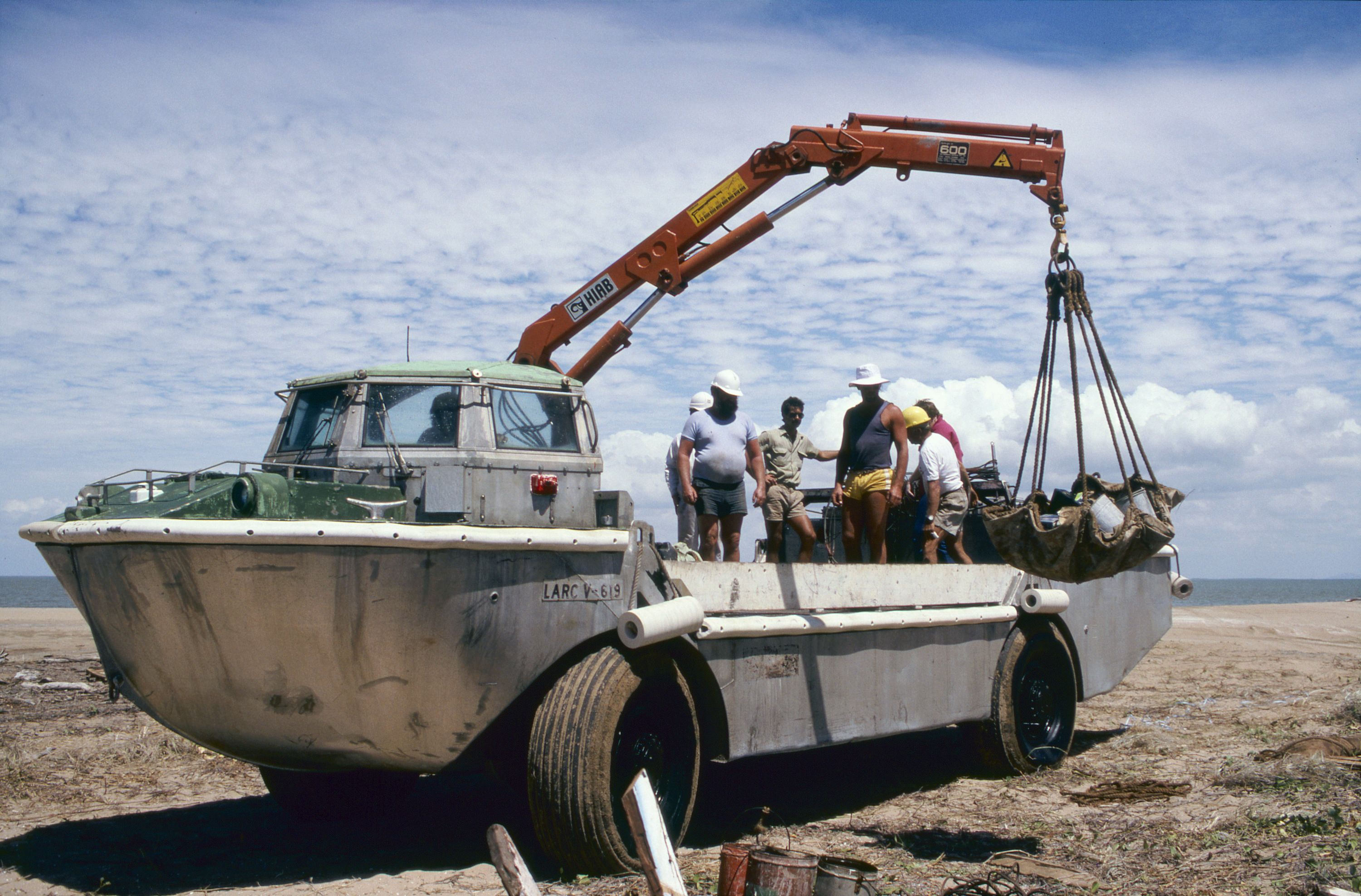 Photo showing an amphibious vehicle is pictured on sandy terrain carrying a team of workers. It is in the process of lifting lighthouse fittings by a crane hoist.