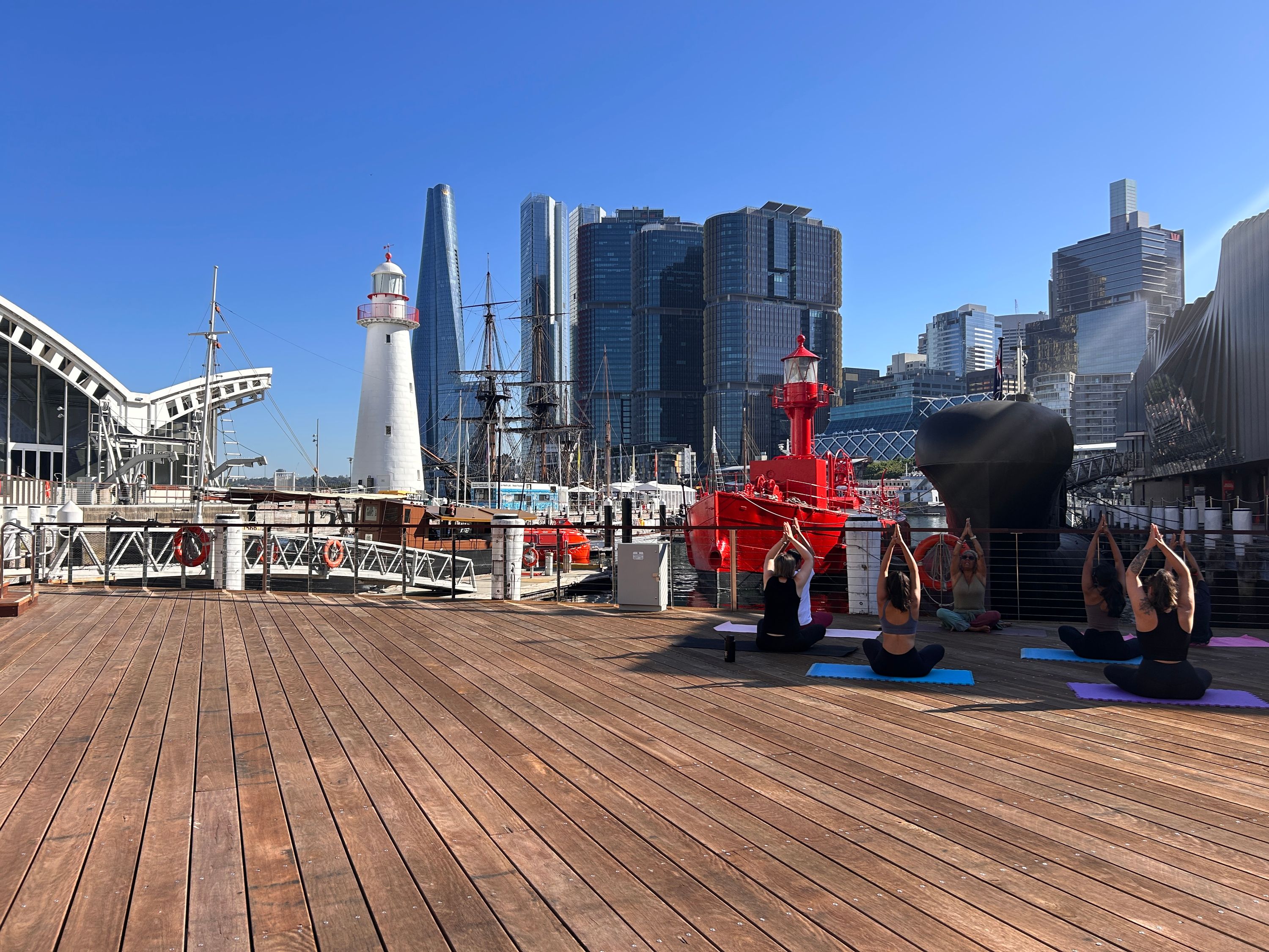 Photograph of the museum waterfront, with a group of people doing yoga on the right hand side. There is a lighthouse on the left and the city buildings in the background.