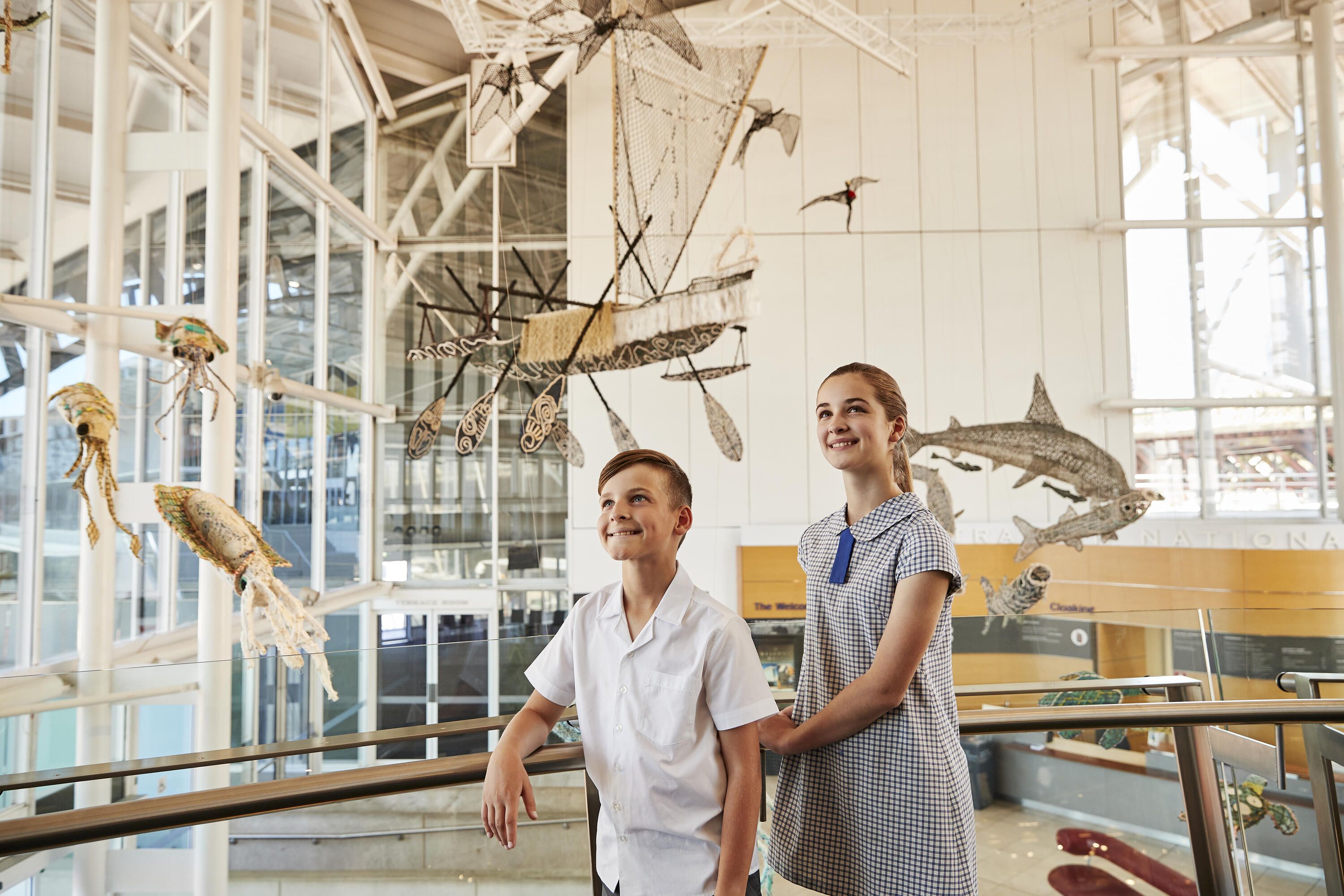 2 School kids exploring the museum, stop to look at the ghost net sculptures suspended in the foyer.