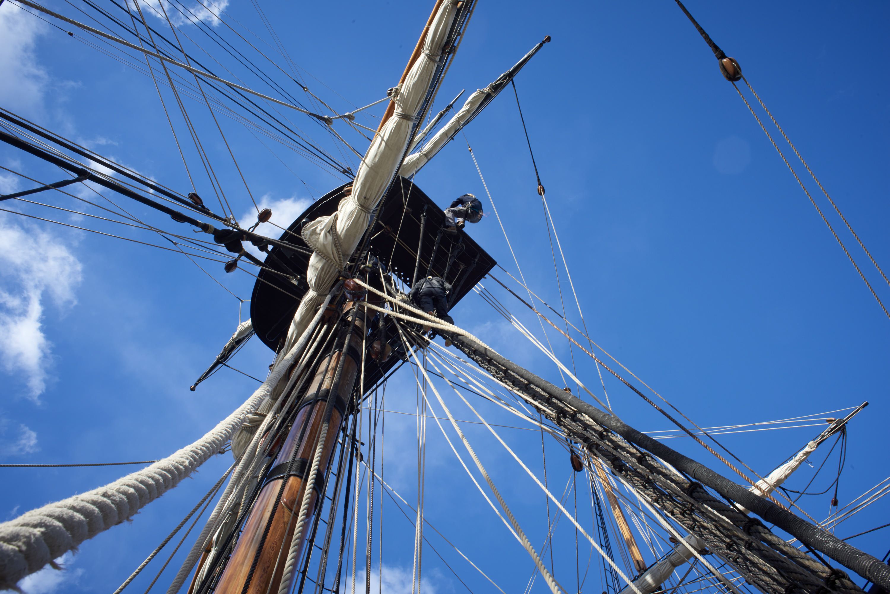 Photo taken looking up at a tall ship mast and rigging with a blue sky.