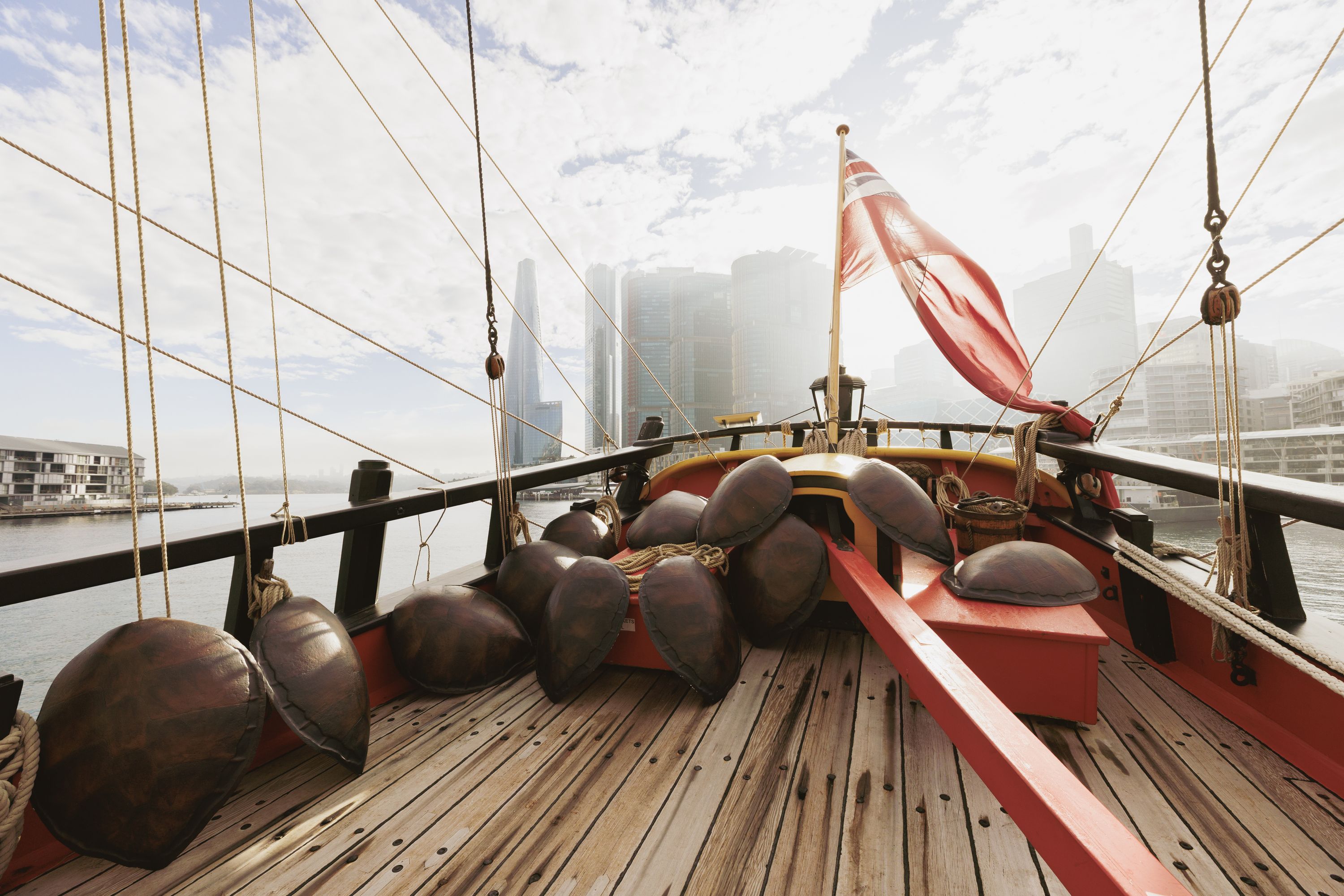 Photo showing the back of a wooden tall ship with a collection of turtle shells on the deck. There is a red flag flying on the ship, and city buildings in the background. 