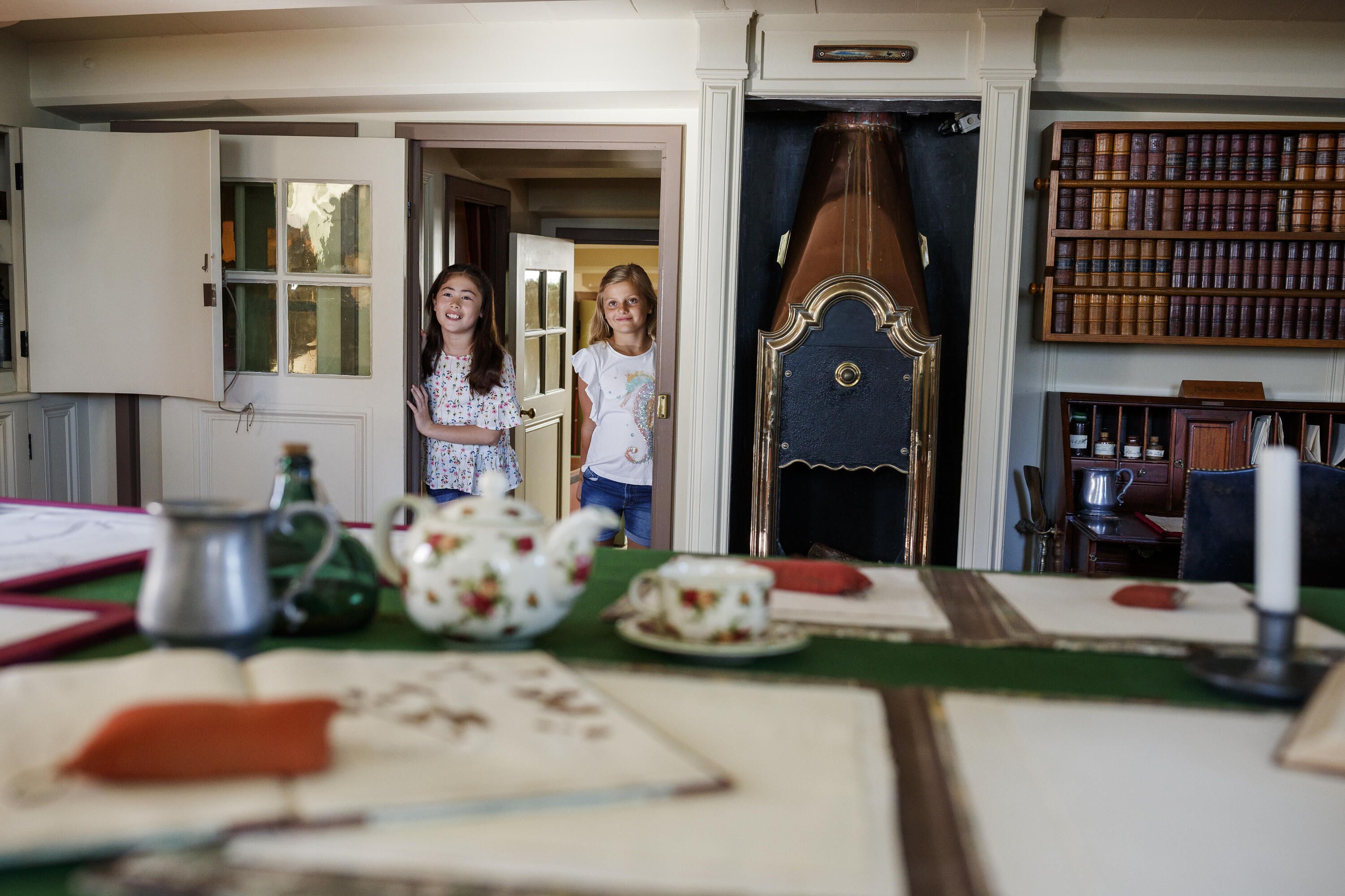 Photo taken inside a tall ship with two girls looking through a doorway towards a table covered in documents and a tea set. There is a fireplace and bookshelf to one side.
