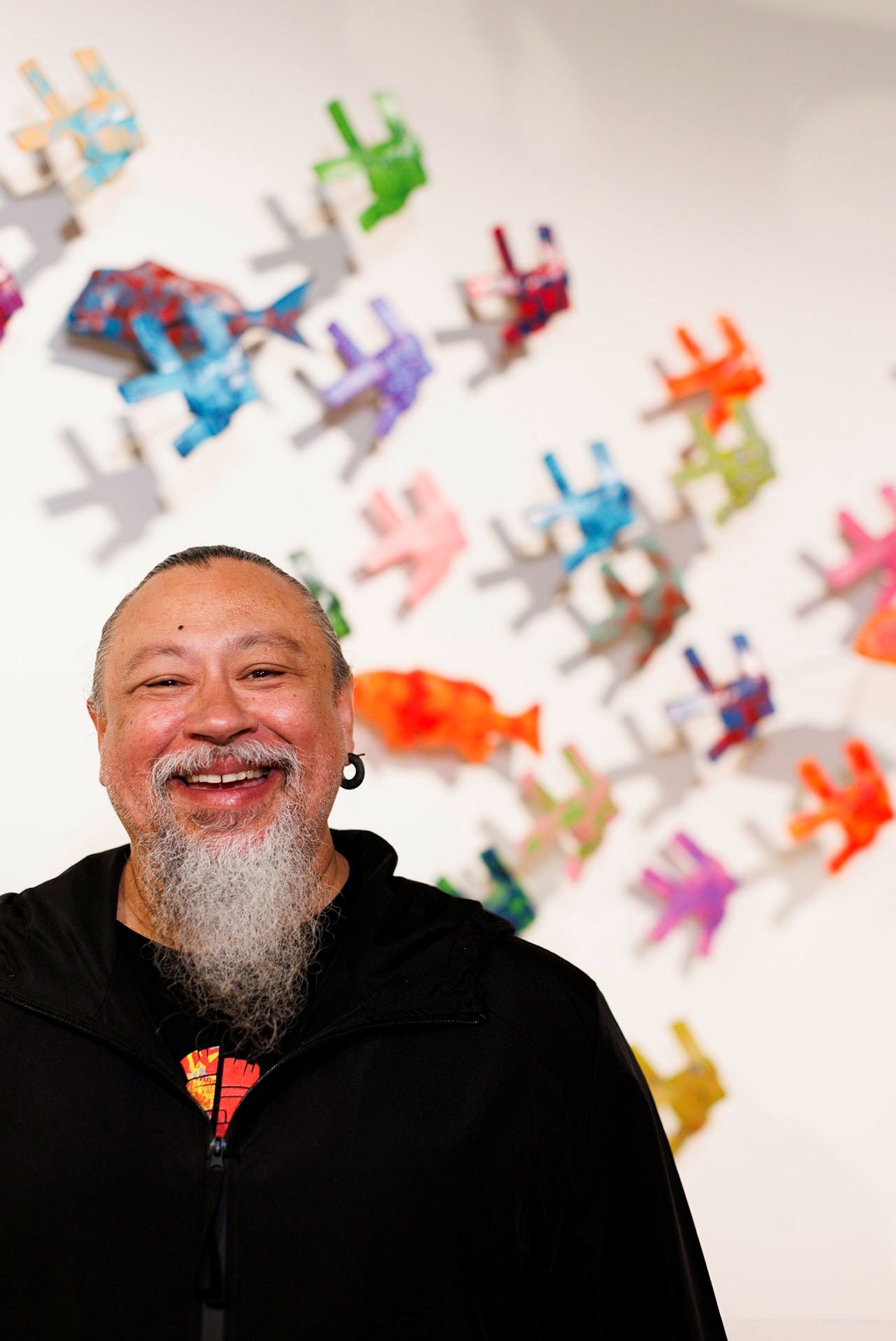 Photo of a smiling man with a grey beard and black hair, standing in front of colourful, woven fish sculptures on a wall. 