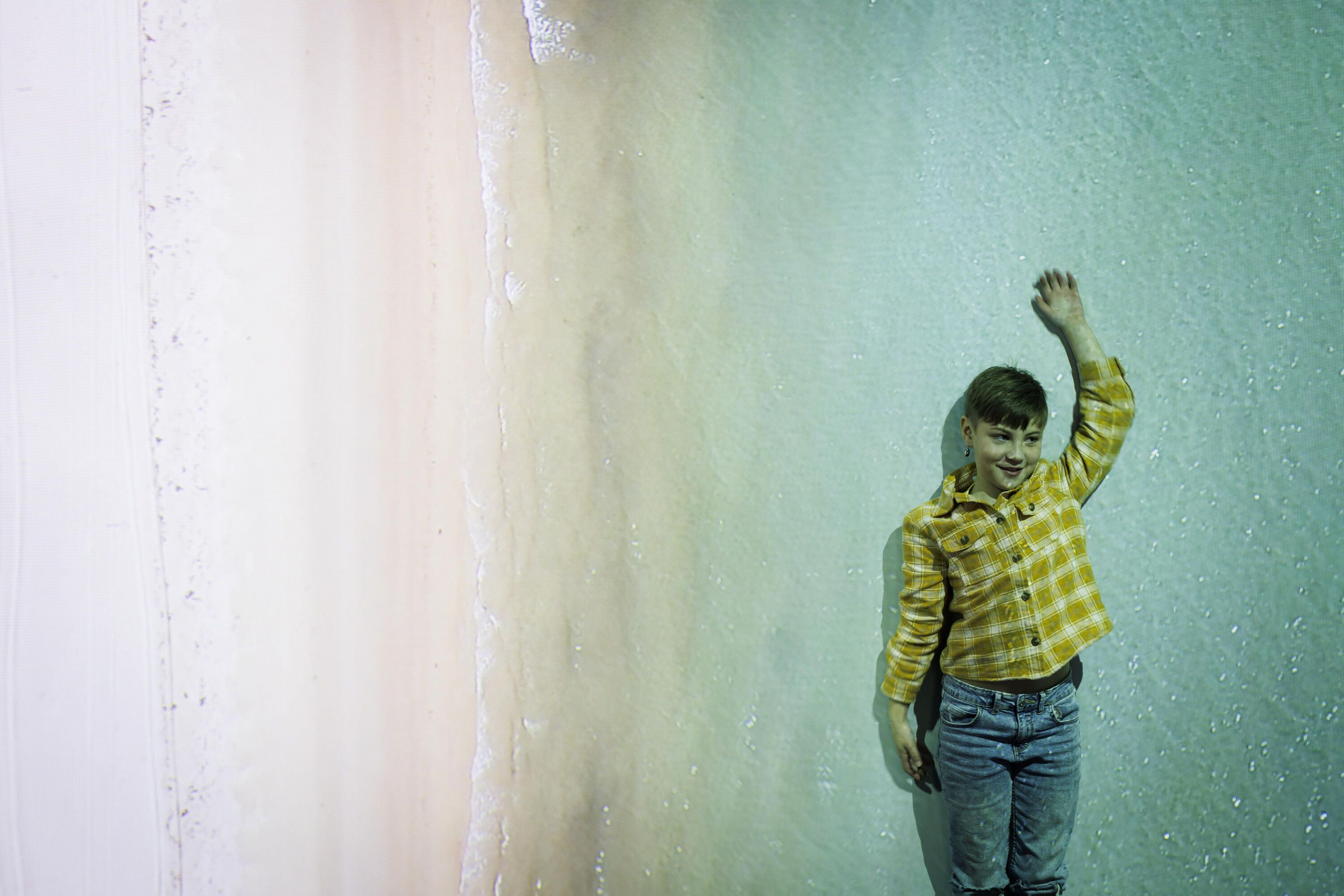 Young boy with short, dark hair, wearing a yellow check shirt standing in front of a large projection of a coastline