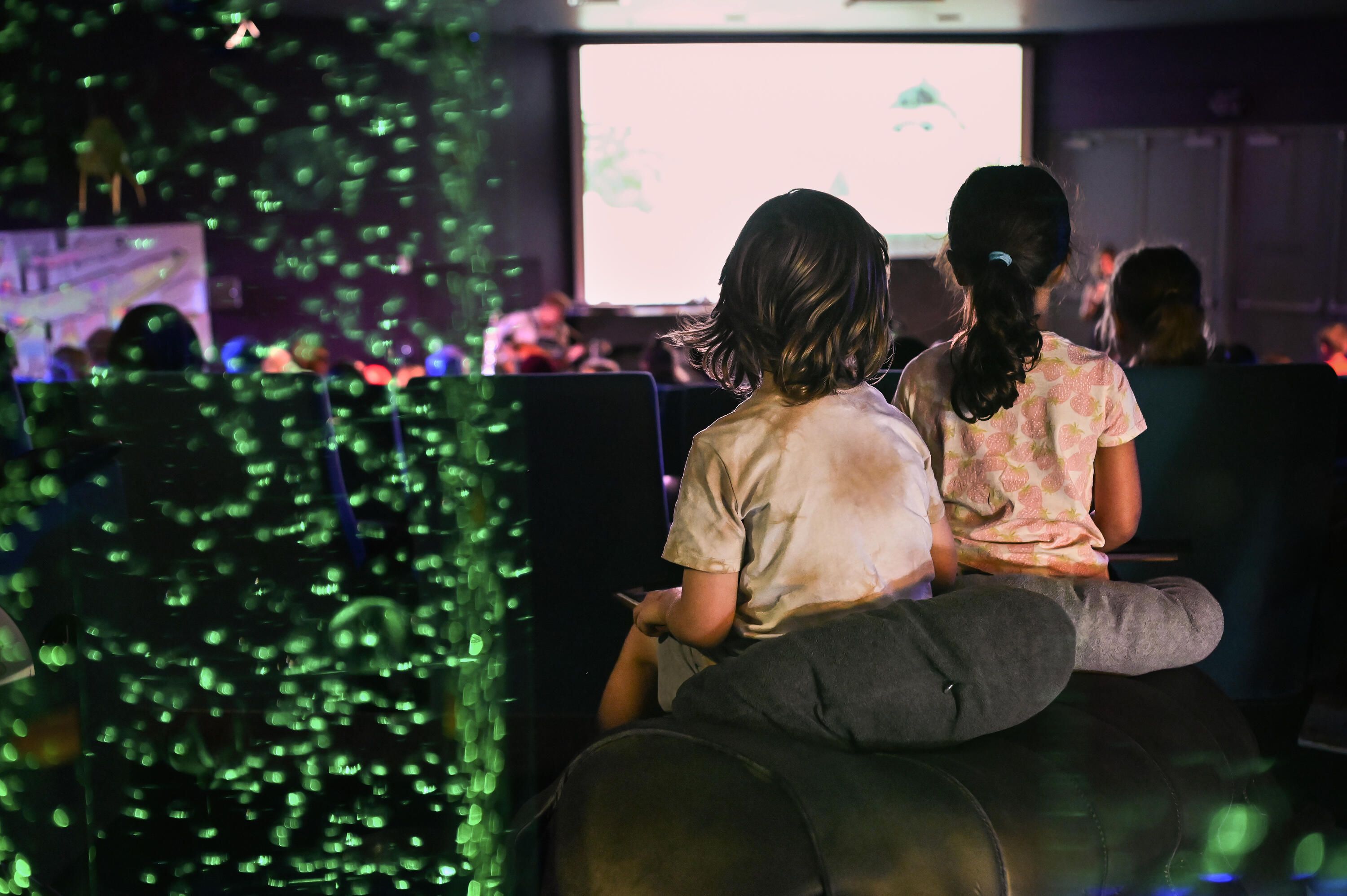 Photo taken from behind showing children in a theatre looking towards the front. There are small green bubbles on the left of the image.