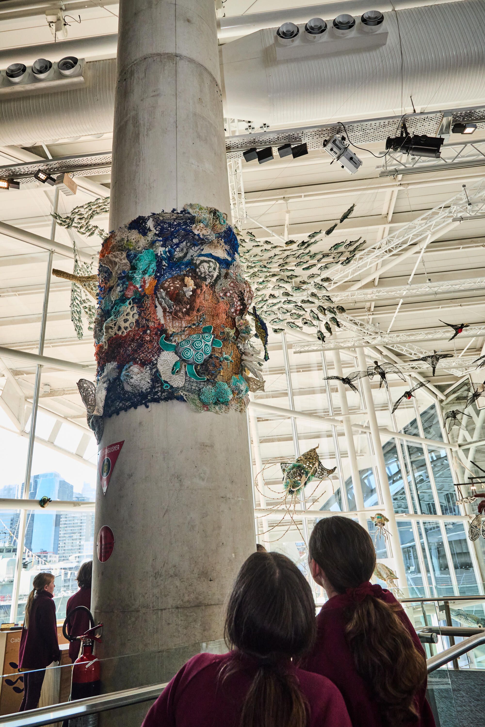 Photo of 2 students looking up at an artwork that looks like a coral reef made from recycled plastic nets.