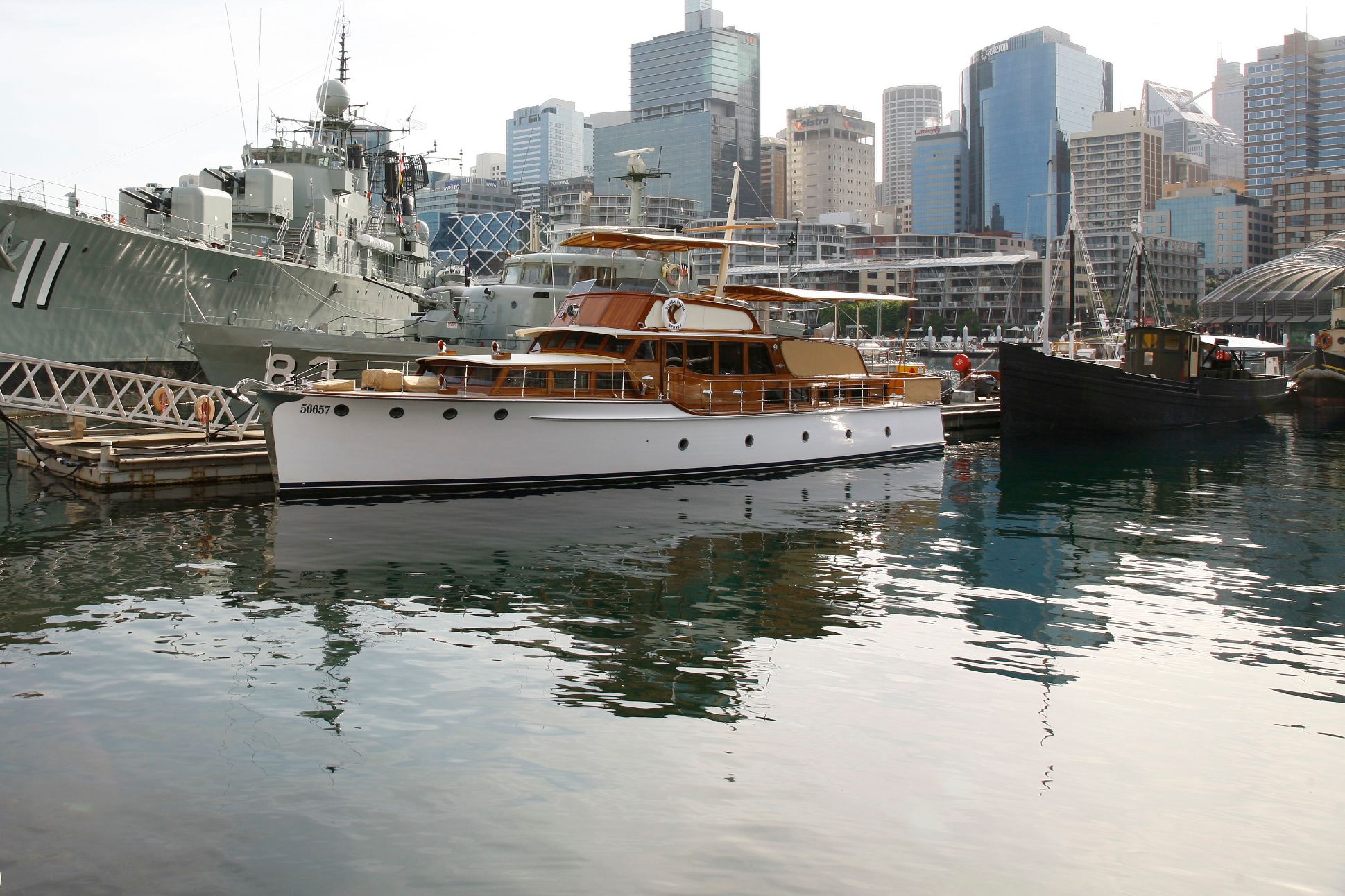 Photo showing a white and wood boat moored at the museum in front of the grey warship vampire. the city buildings are in the background. 