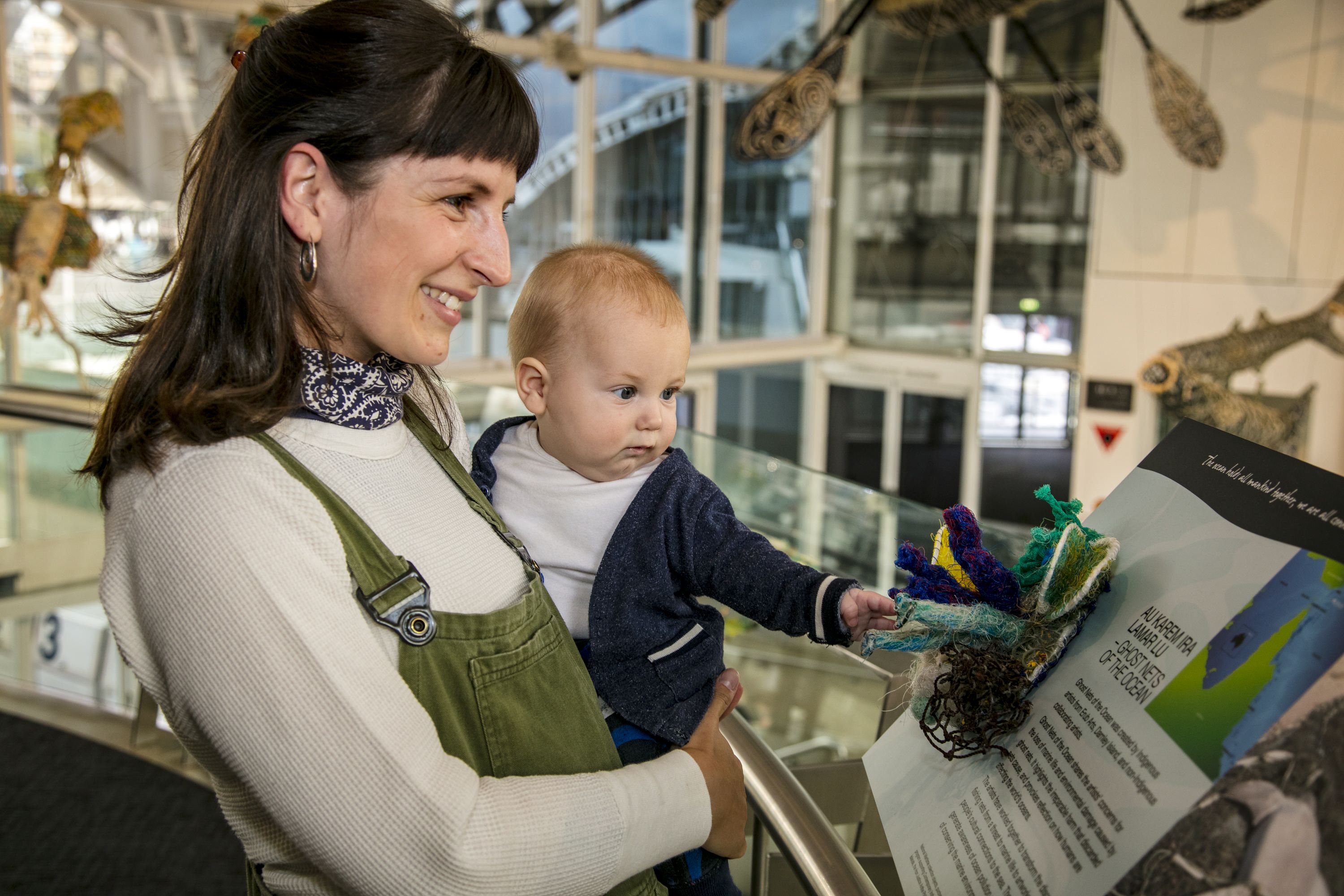 A new young mum, posed holding her baby in the museum with the Ghost Nets foyer display in the background.