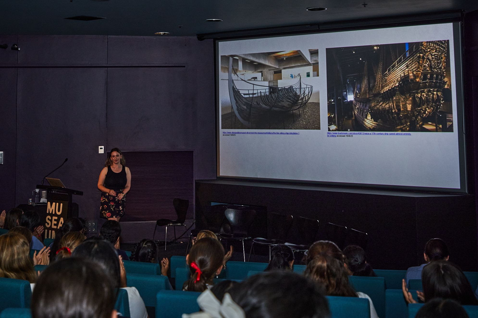 Photo of a theatre looking over the heads of school students at a woman presenting. On the big screen is a presentation with 2 photos of shipwrecked boats in museum exhibitions.