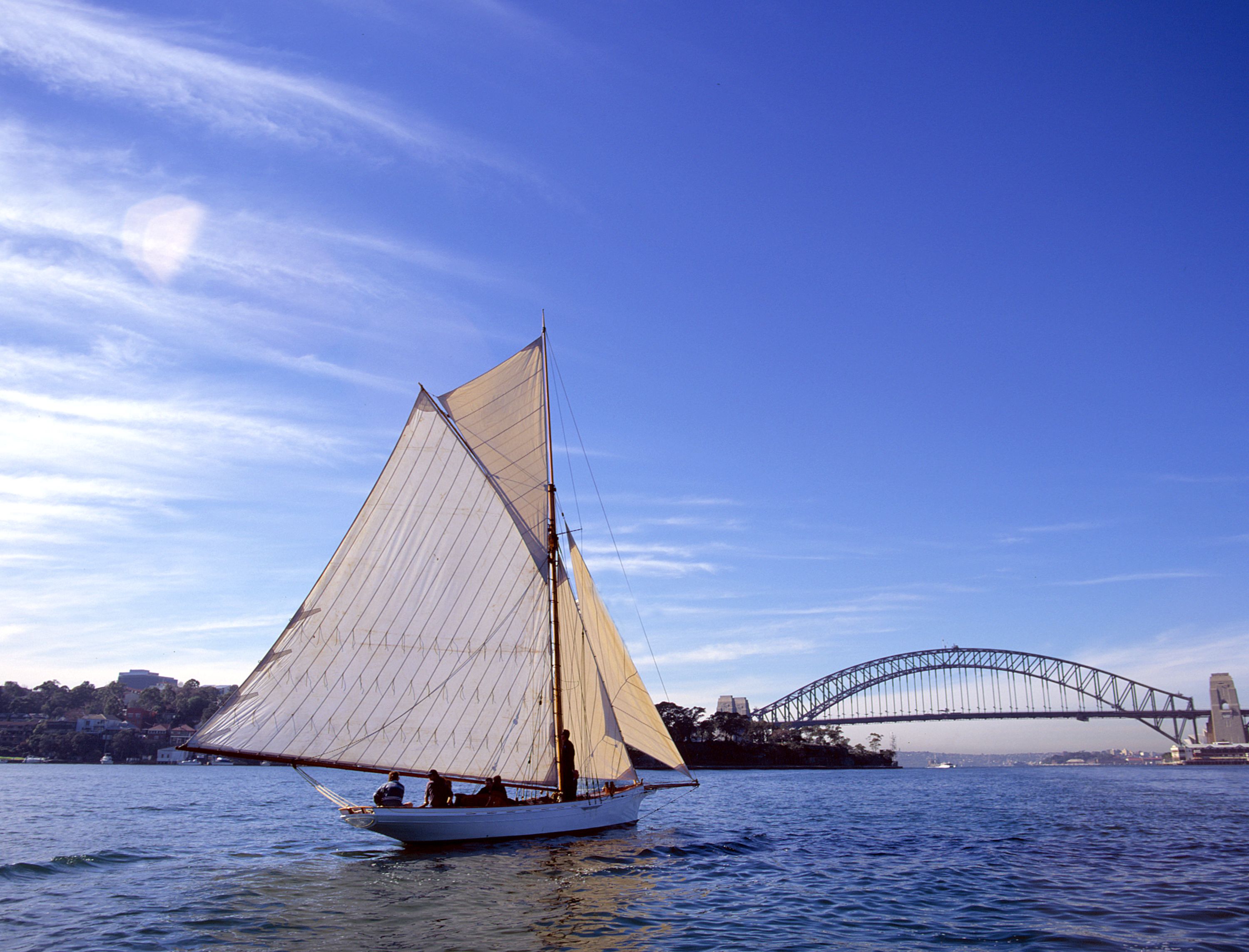 Photo of a small sailing boat, with large white sails on a clear, sunny day. The Sydney harbour bridge is in the background.