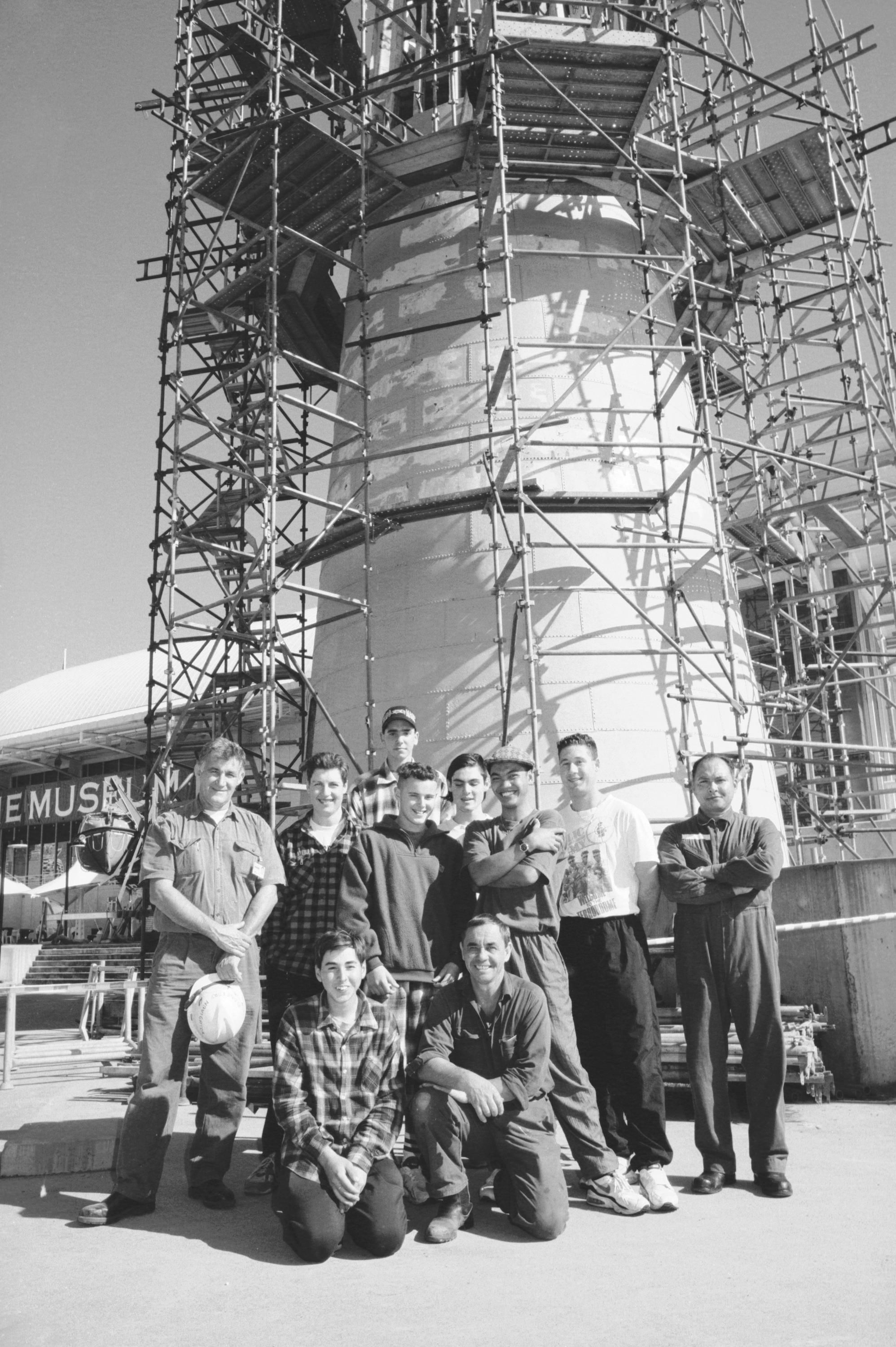 Black and white photo showing a group of men standing in front of a lighthouse while it is under construction. 
