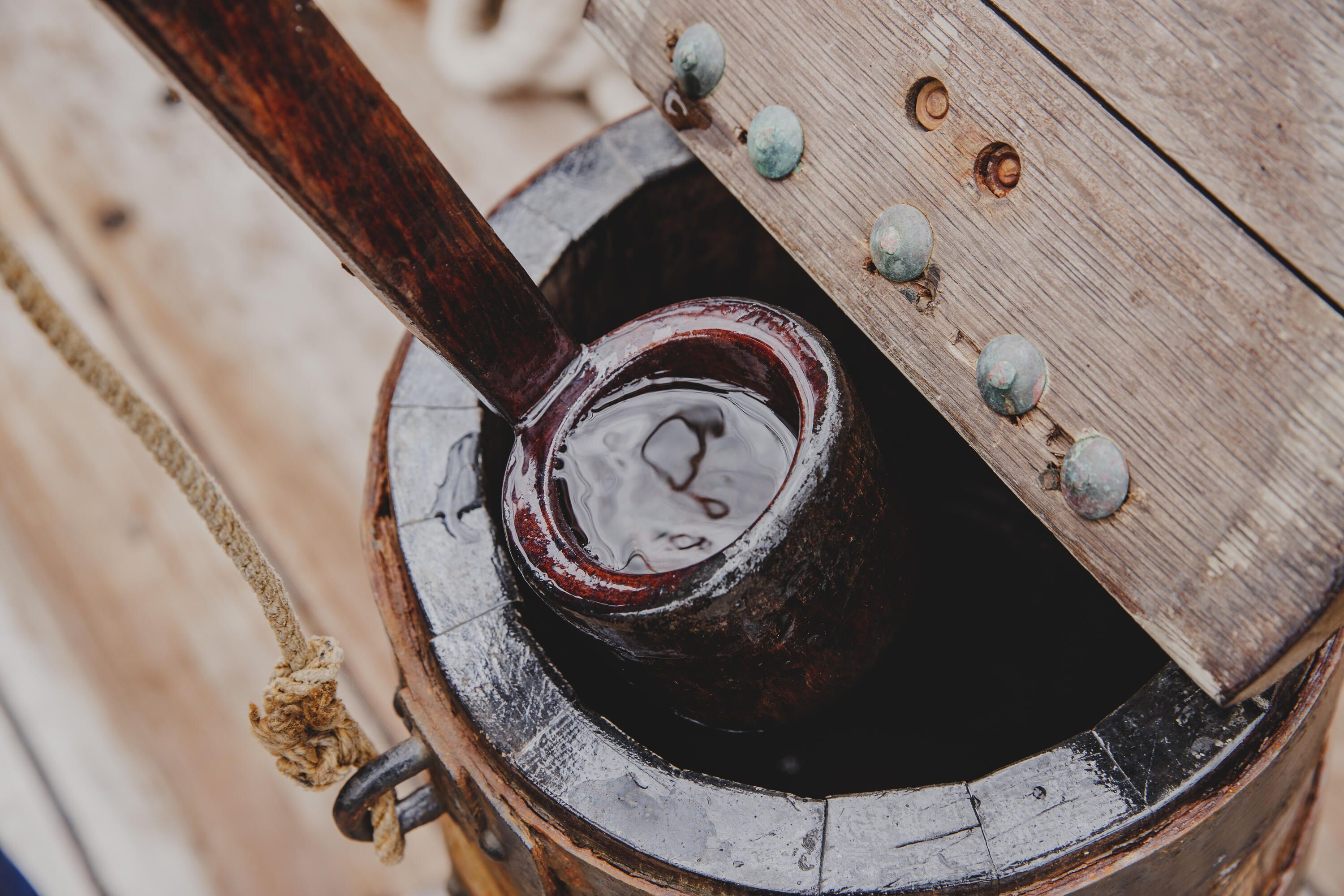Close up photo showing a wooden dipper lifting water out of a lidded barrel on the upper deck of a wooden tall ship.