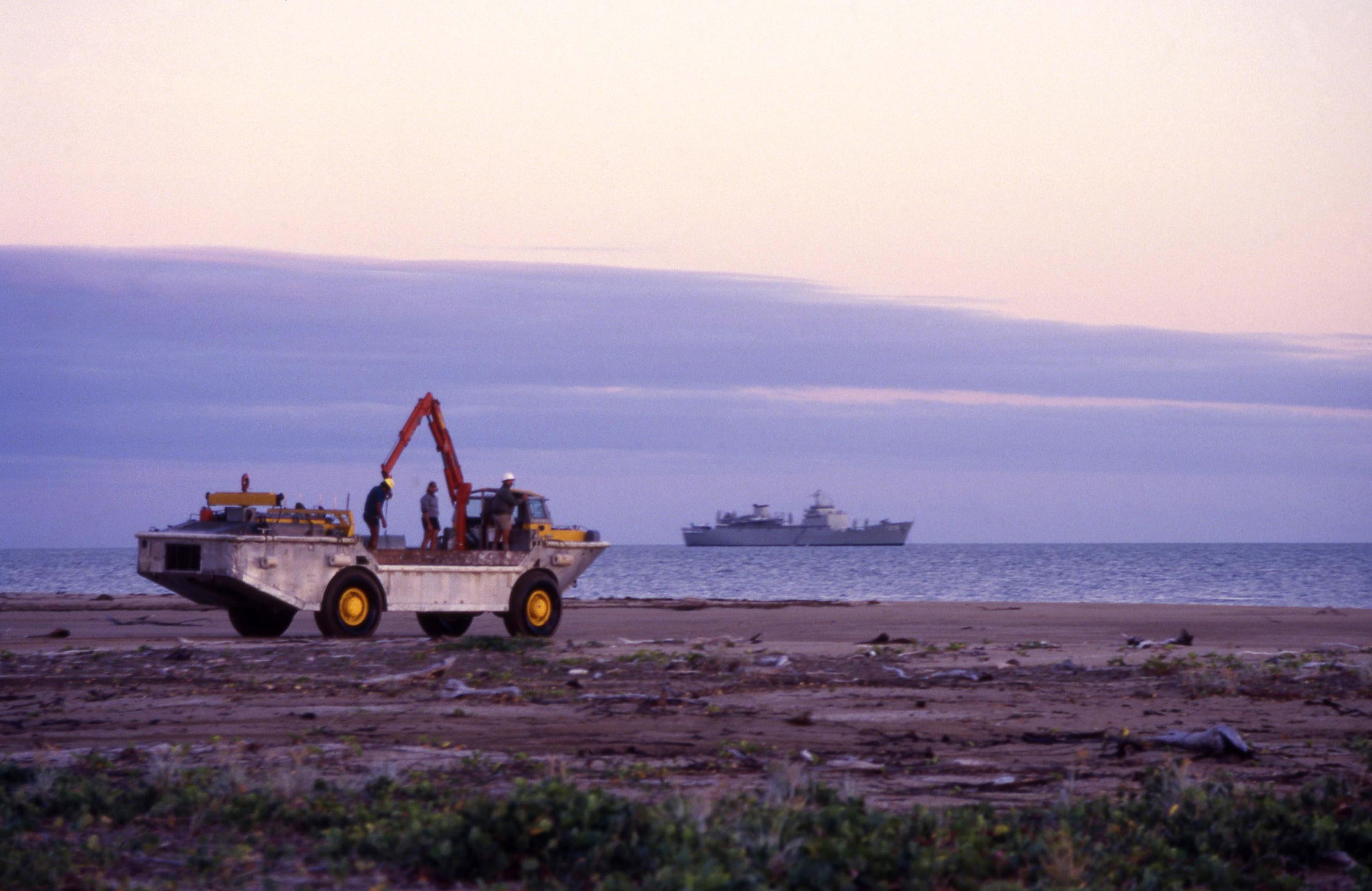 Photo of an amphibious vehicle driving along a beach, a navy ship is on the water in the distance.