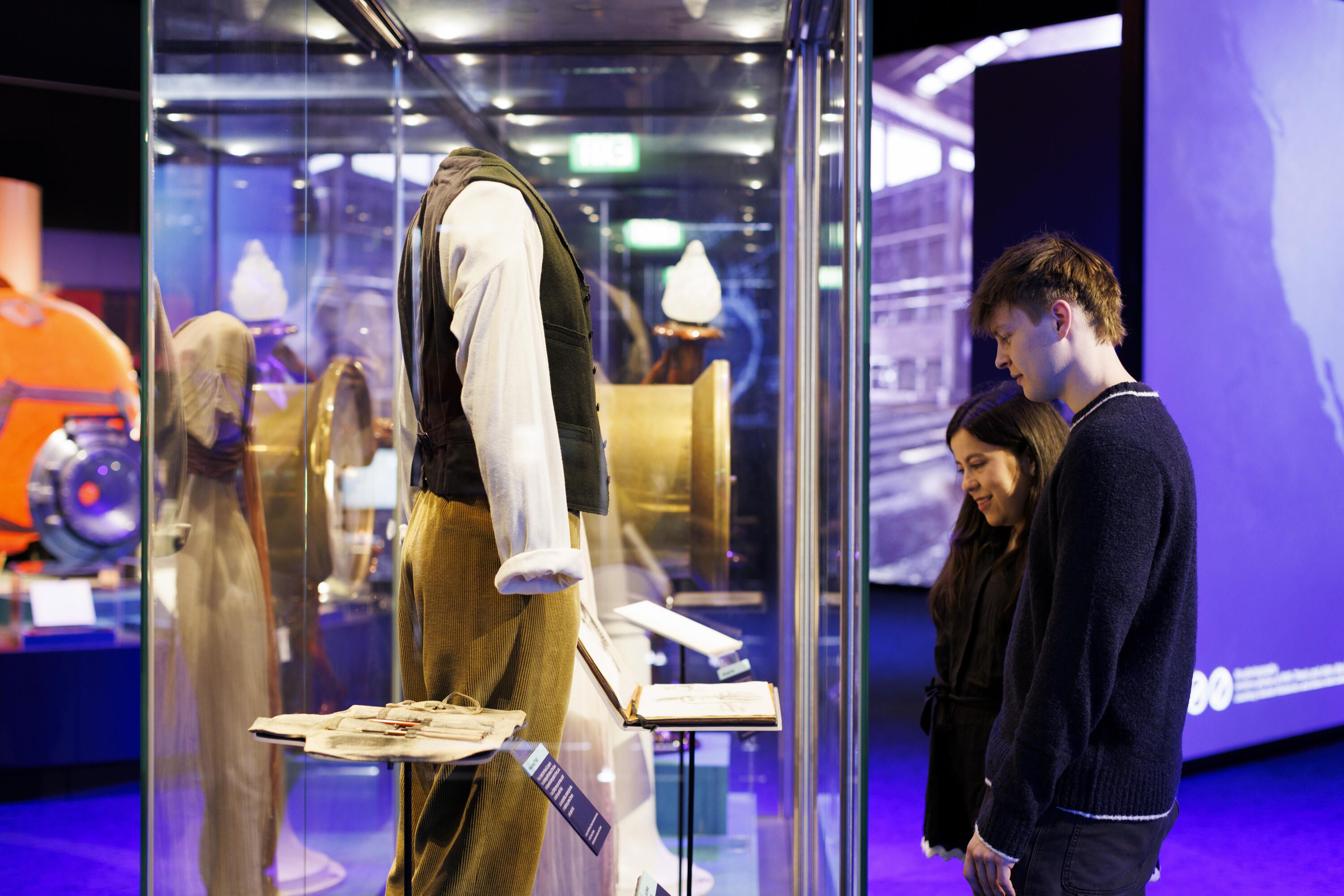 A man and a woman look at a museum case containing costumes and props from the movie Titanic. 