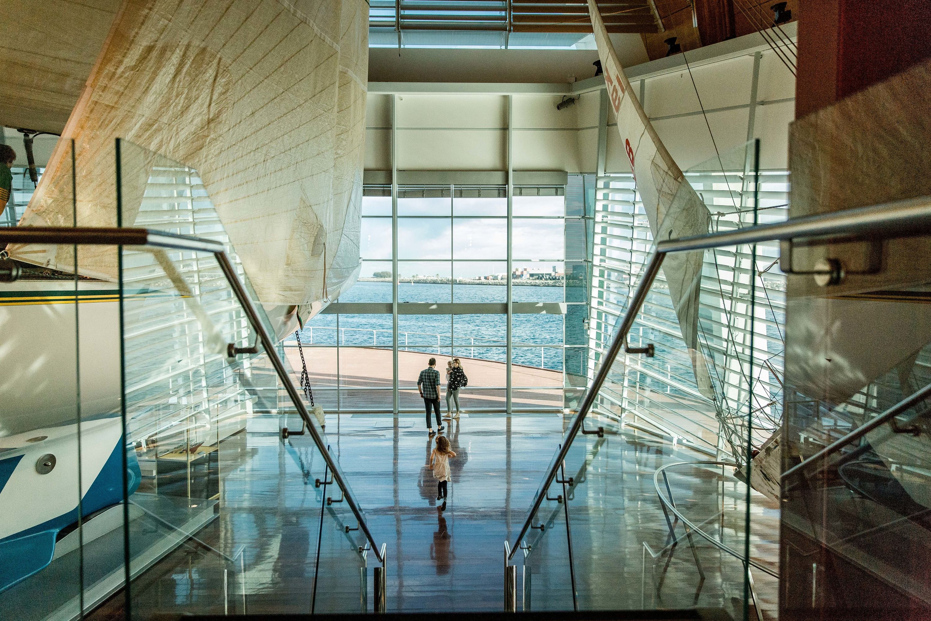 Photo of the inside of a large museum space containing several boats, looking out a window to the harbout 