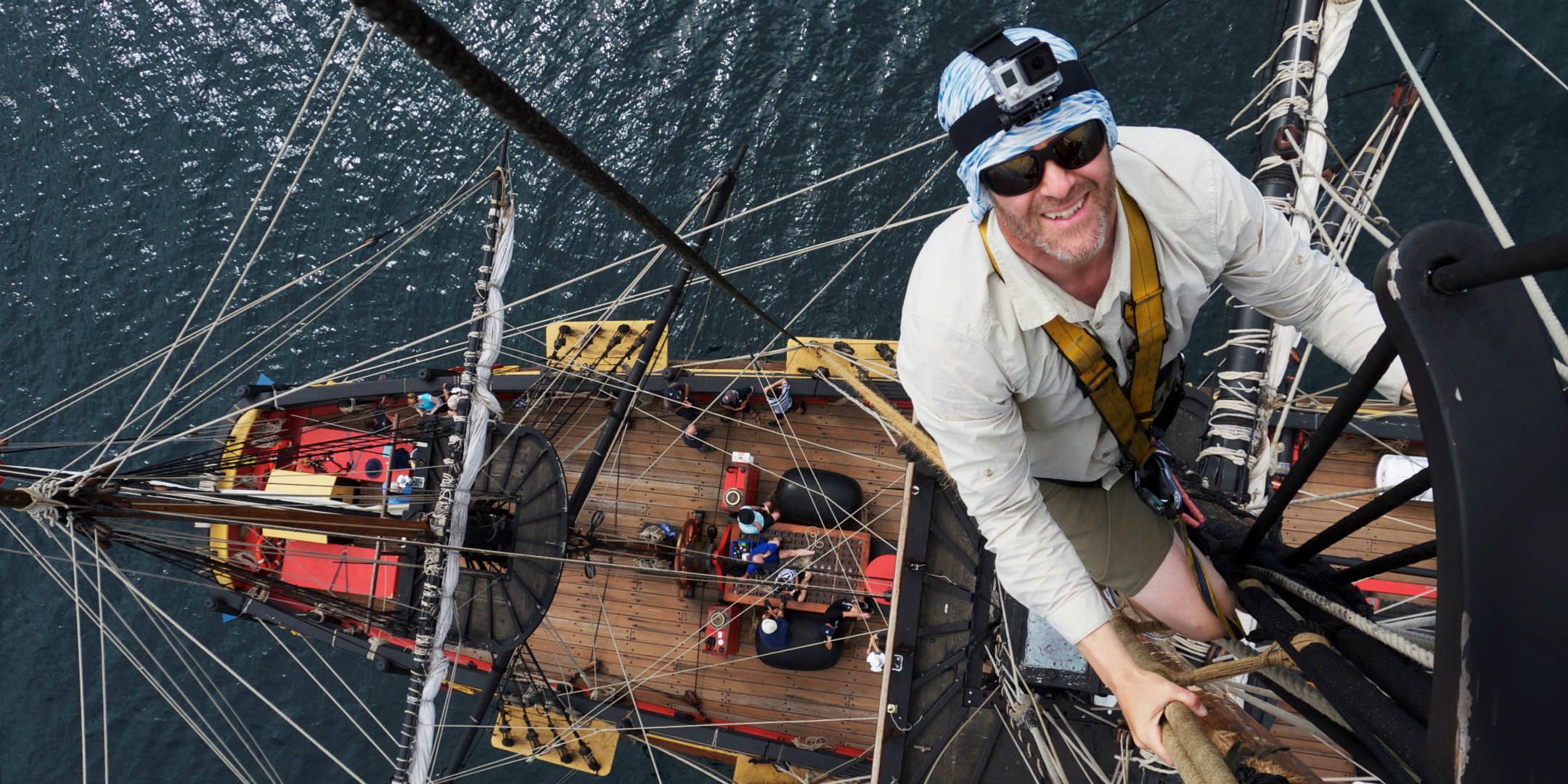 Photo taken from above of a man wearing sunglasses and a helmet climbing the rigging of endeavour. He is high up, wearing a harness connected to the ship, and you can see the deck of the ship and the ocean below.