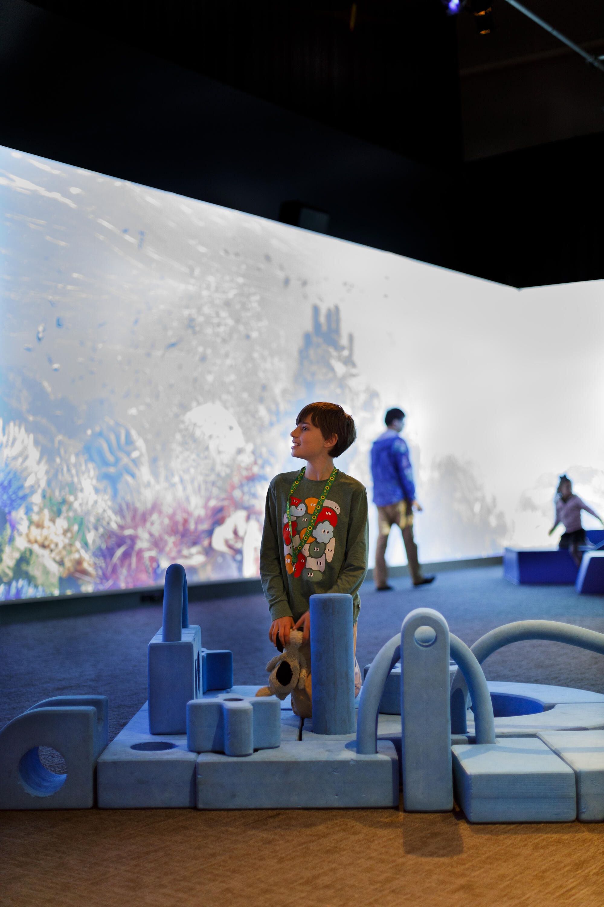 Young boy with short, dark hair, wearing a grey shirt with colourful print and sunflower lanyard playing with large blue blocks.