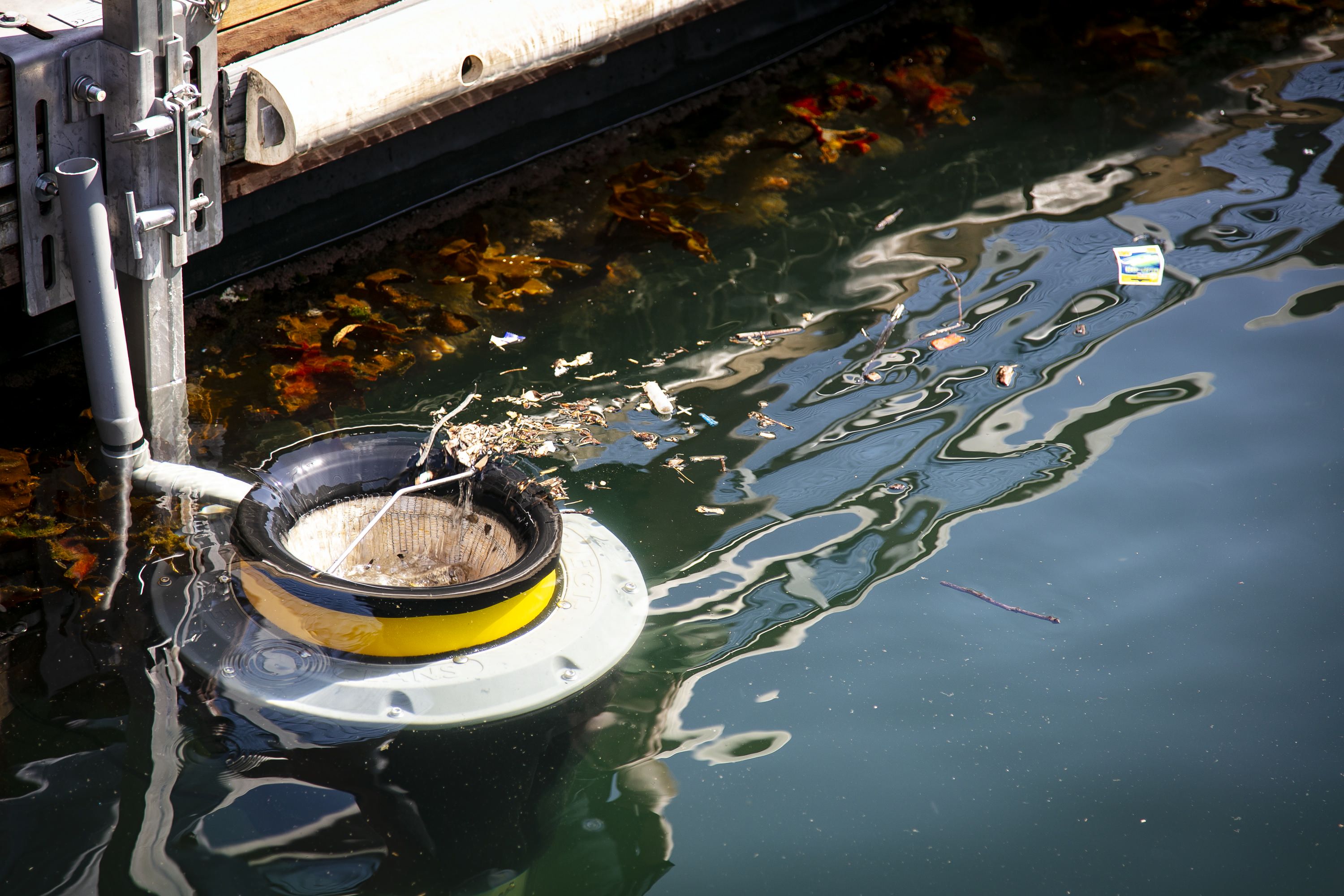 Photo showing a yellow 'bin' in the water attached to a floating wharf. Rubish and leaves are floating towards the bin.