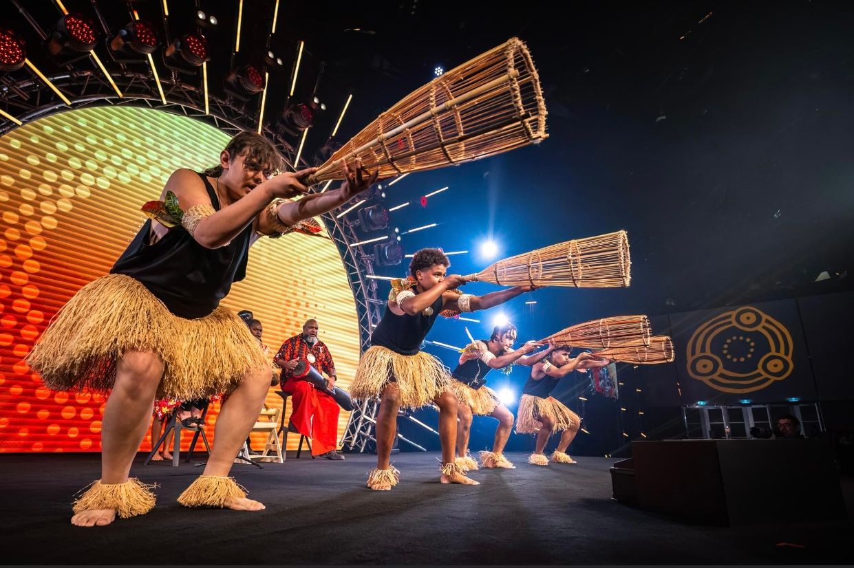 Photo of dancers wearing black tops and grass skirts on a stage with an orange and yellow sun set behind them.