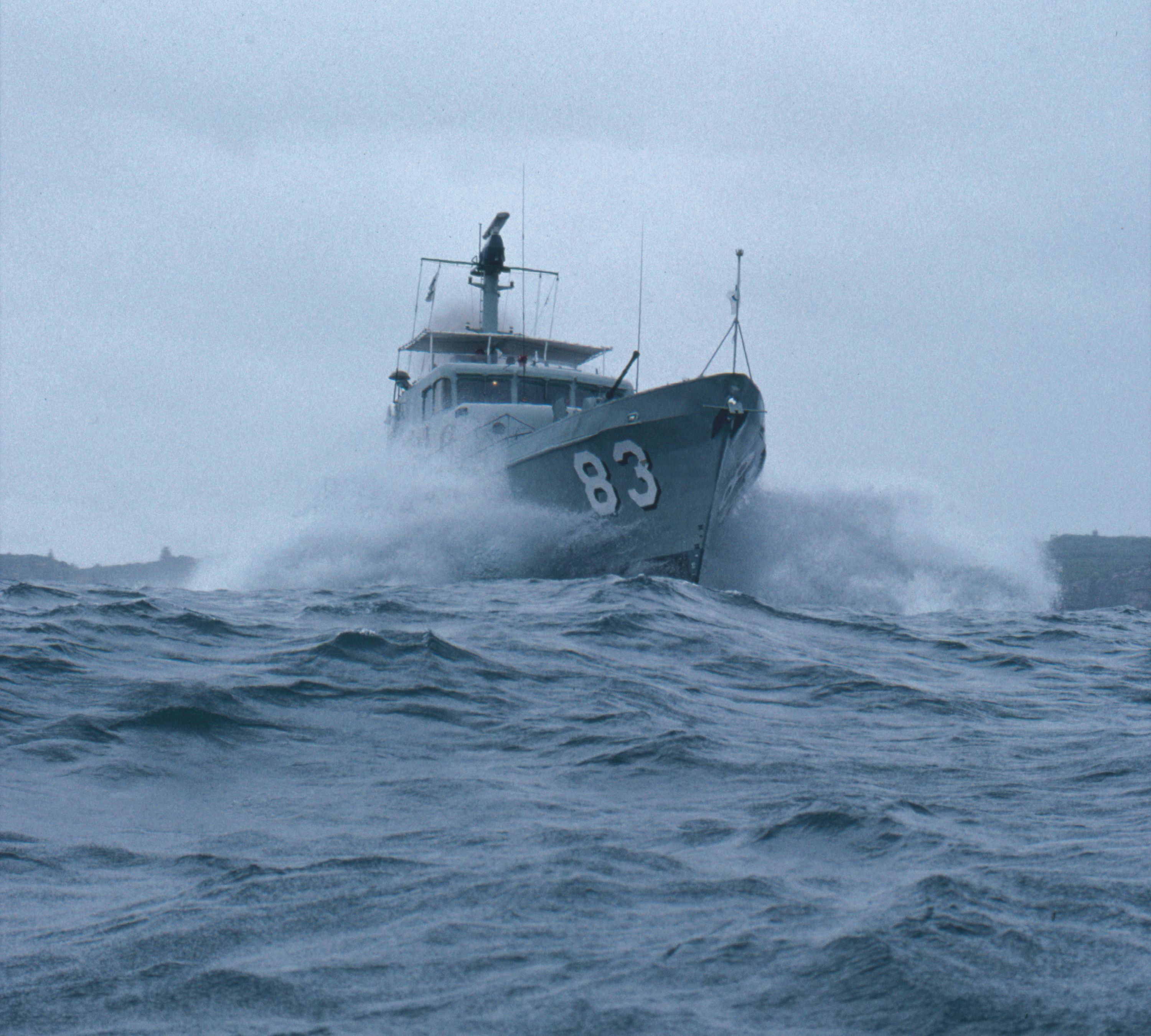 Photo of a small, grey boat crashing through the waves, with the number 83 on its bow. 