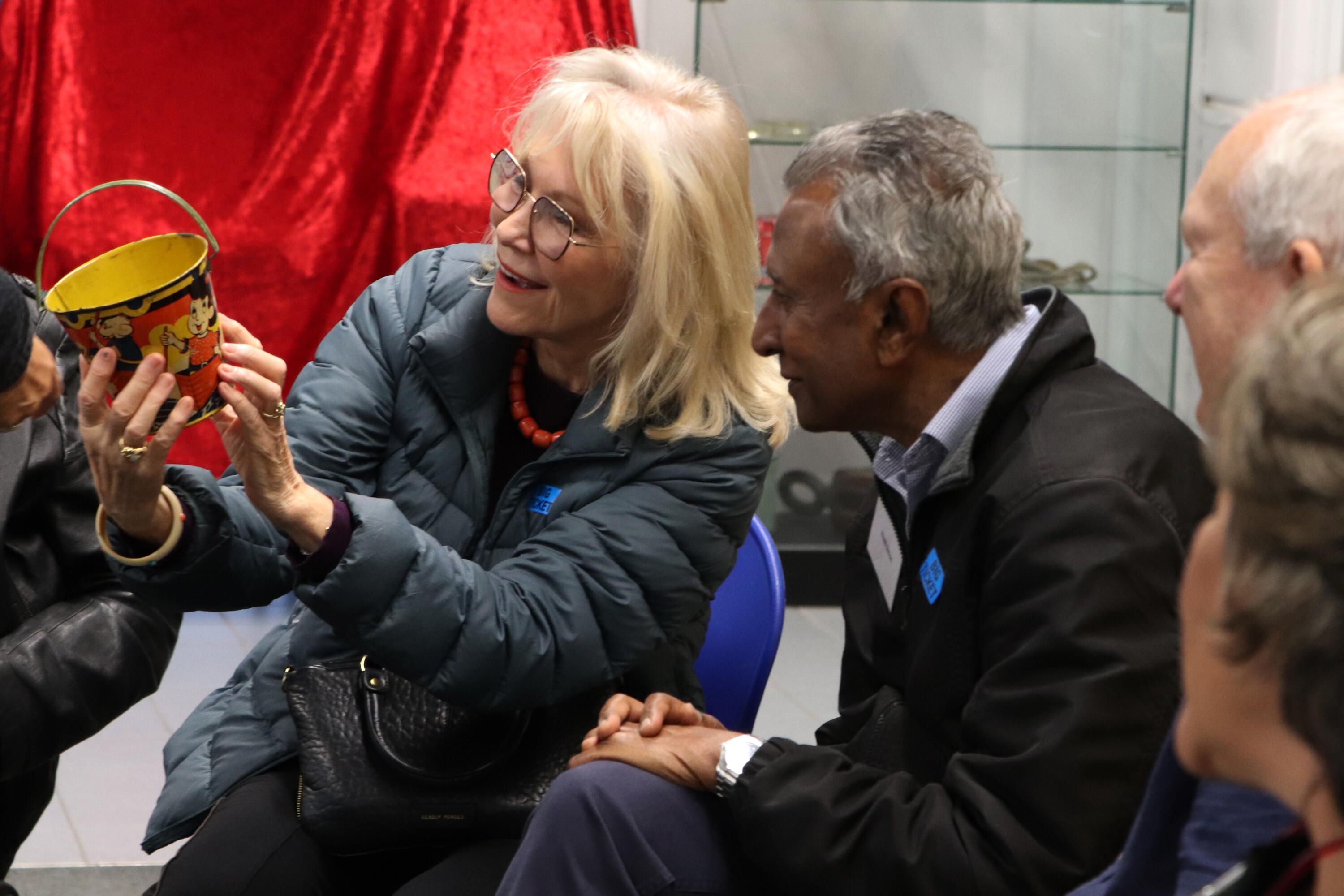 Lady with blonde hair and glasses holding a metal bucket and showing it to a man with dark skin and grey hair.